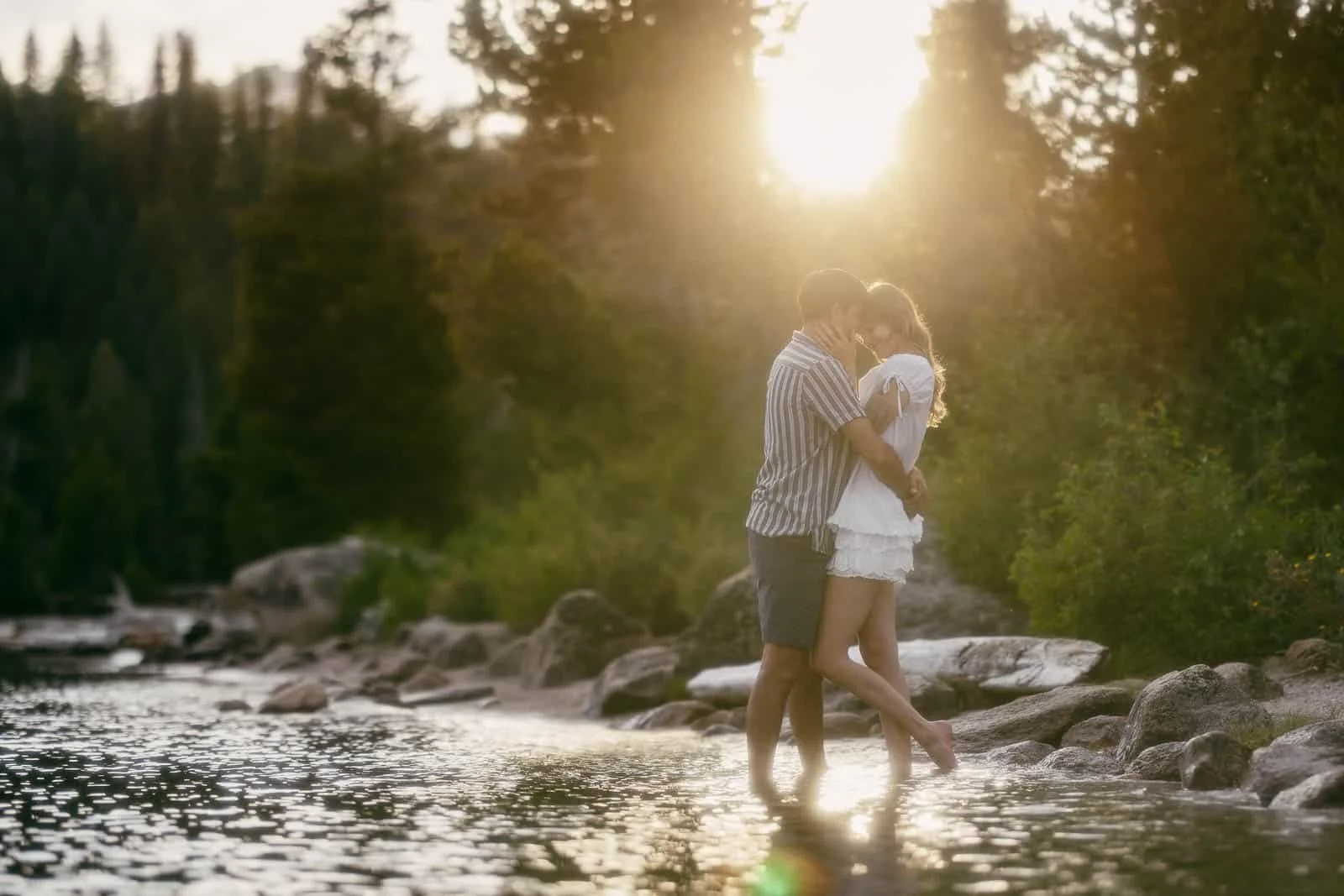  A couple stands in shallow water by a lake, embracing at sunset. Sunlight filters through the trees, creating a warm, romantic atmosphere. 