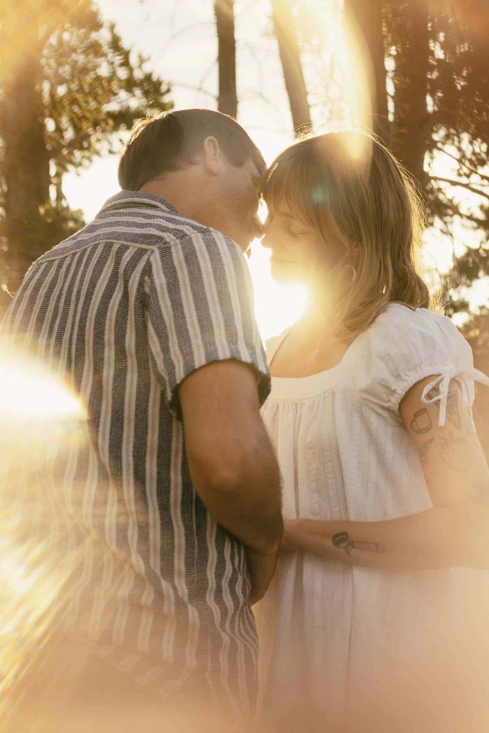  A couple stands close together in a sunlit forest, touching foreheads and holding hands. The sunlight creates a warm, glowing effect around them. 