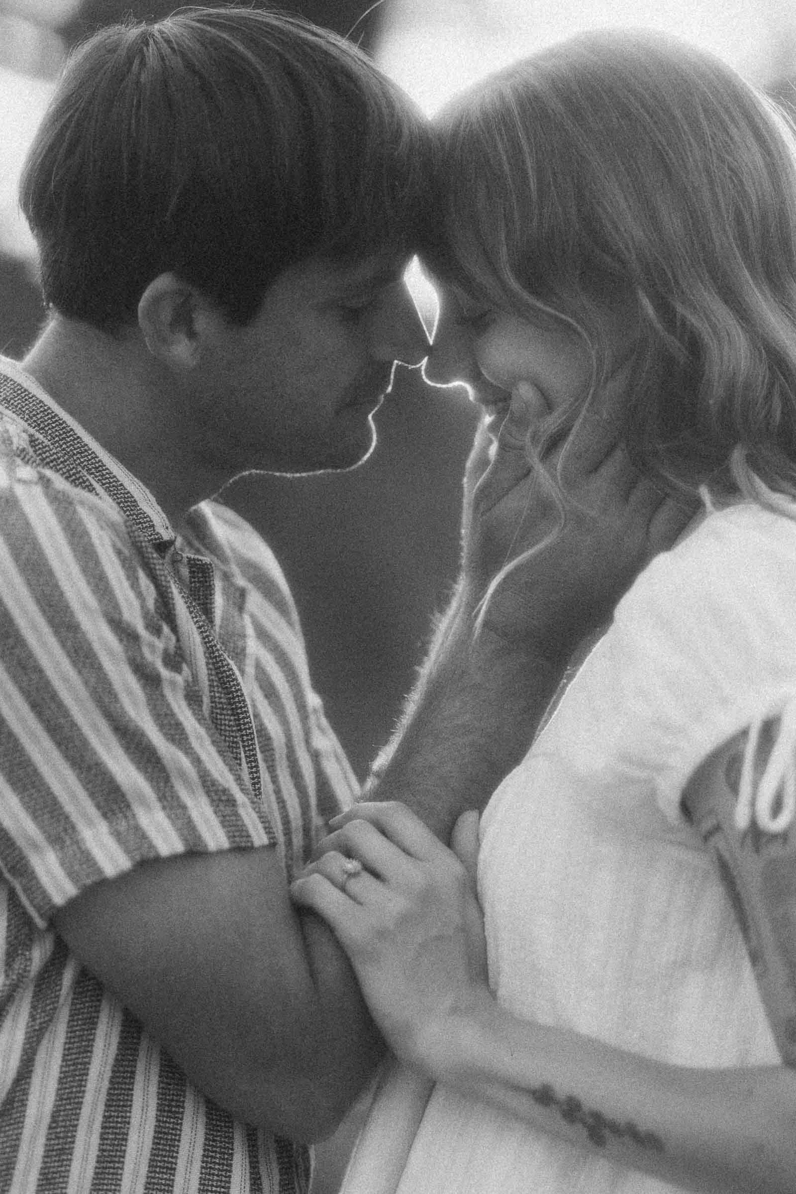  A couple stands closely, touching foreheads and smiling softly. The man gently holds the woman’s face, and she rests her hand on his chest. The image is black and white with a soft, romantic focus. 