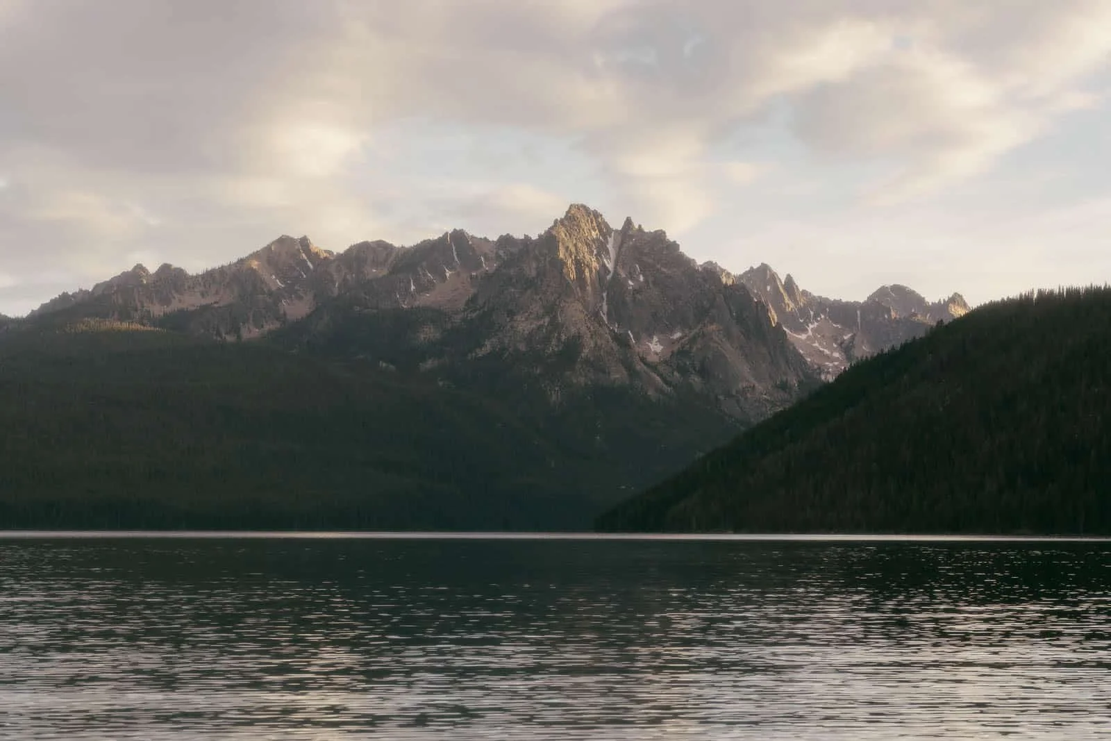  Redfish Lake reflects distant pine-covered mountains under a cloudy sky, with sunlight softly illuminating the rugged mountain peaks. 