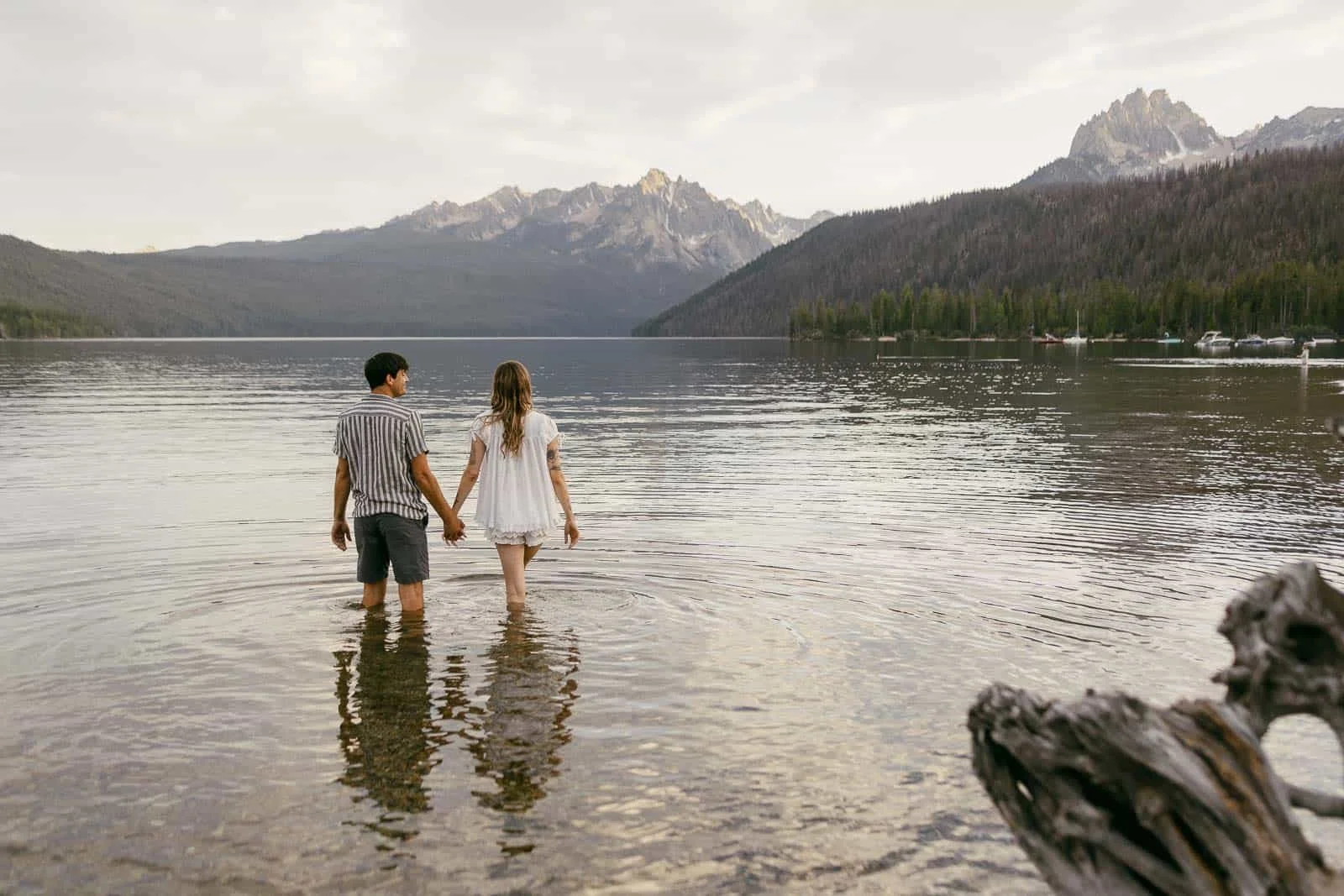  A couple stands hand in hand in a lake, facing distant mountains and a forested shoreline, with gentle ripples in the water around them. 