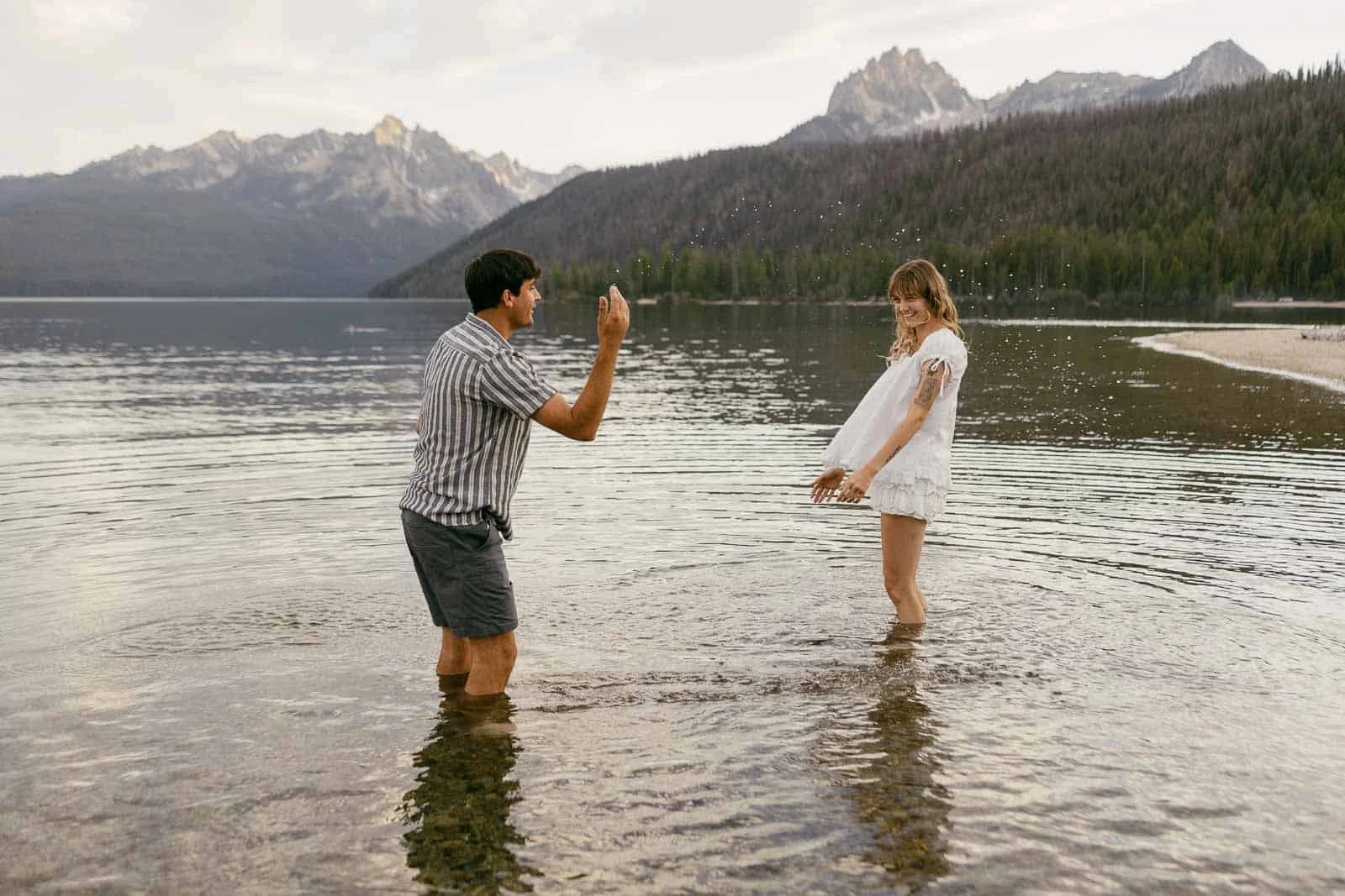  A man and woman playfully splash water at each other while standing in a shallow lake, surrounded by mountains and forest under a cloudy sky. 