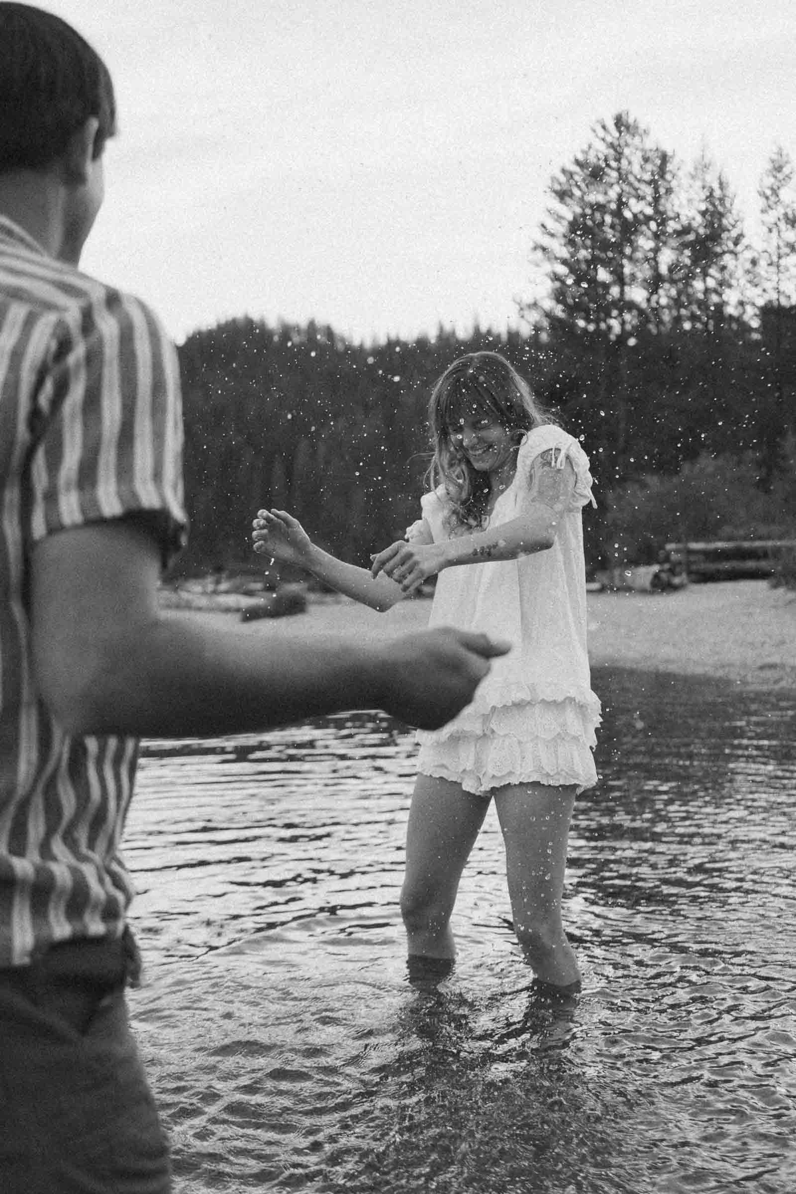  A woman in a white dress splashes water playfully while standing in a lake, smiling. A man in a striped shirt stands nearby. Trees and a sandy shore are in the background. 