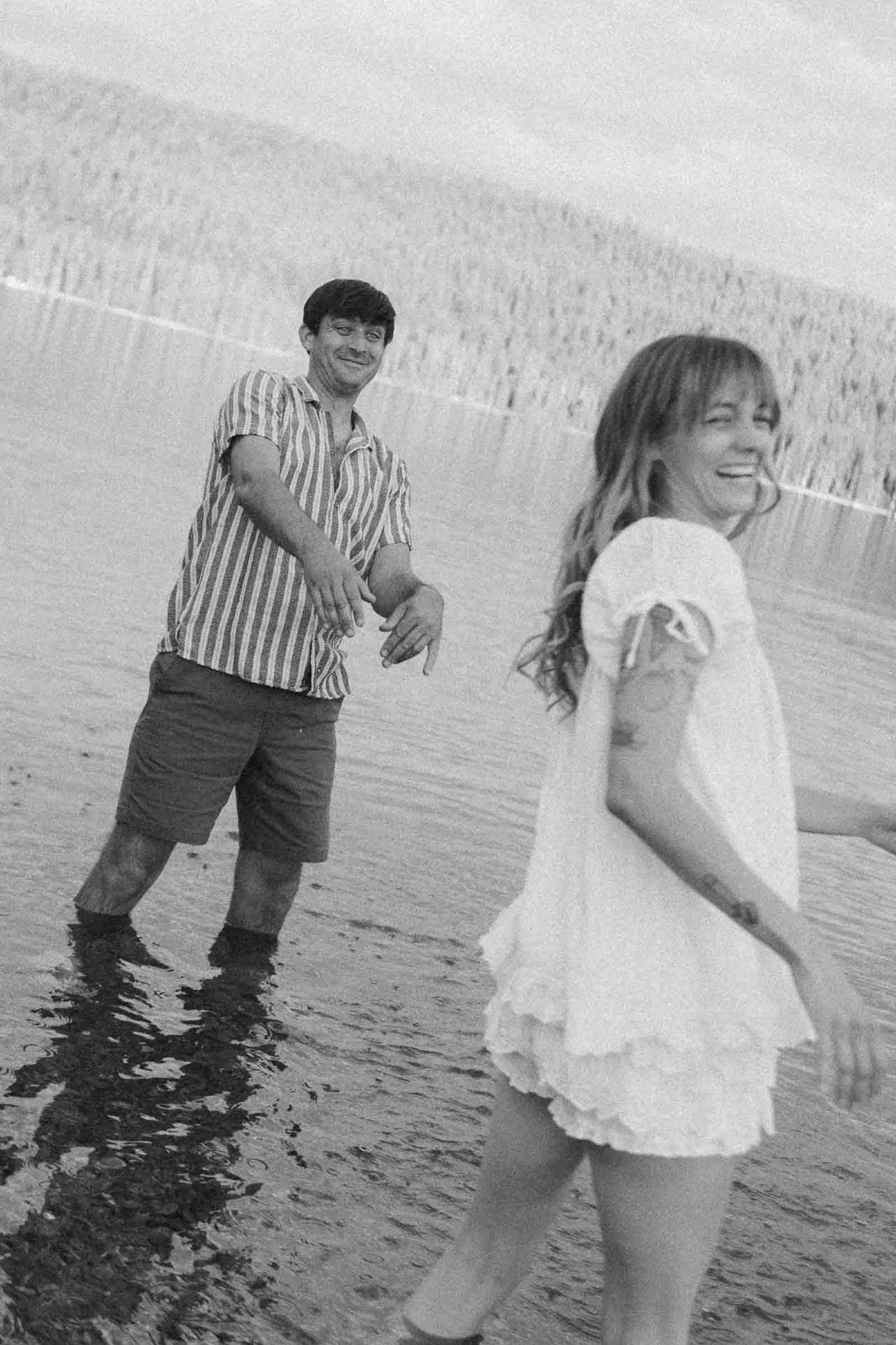 Two people stand in shallow water, smiling and playing. The man wears a striped shirt and shorts, while the woman wears a white dress. Trees and water are visible in the background. The image is in black and white. 