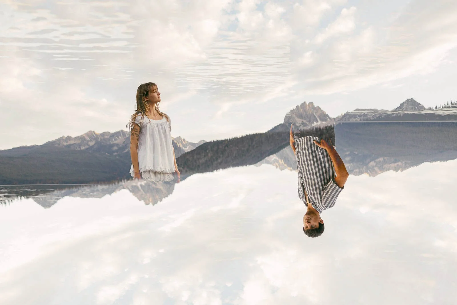 A double-exposed portrait of a woman and a man at the edge of Redfish Lake, one frame flipped upside down over the other so the couple appears to merge into the Sawtooth Mountains behind them.