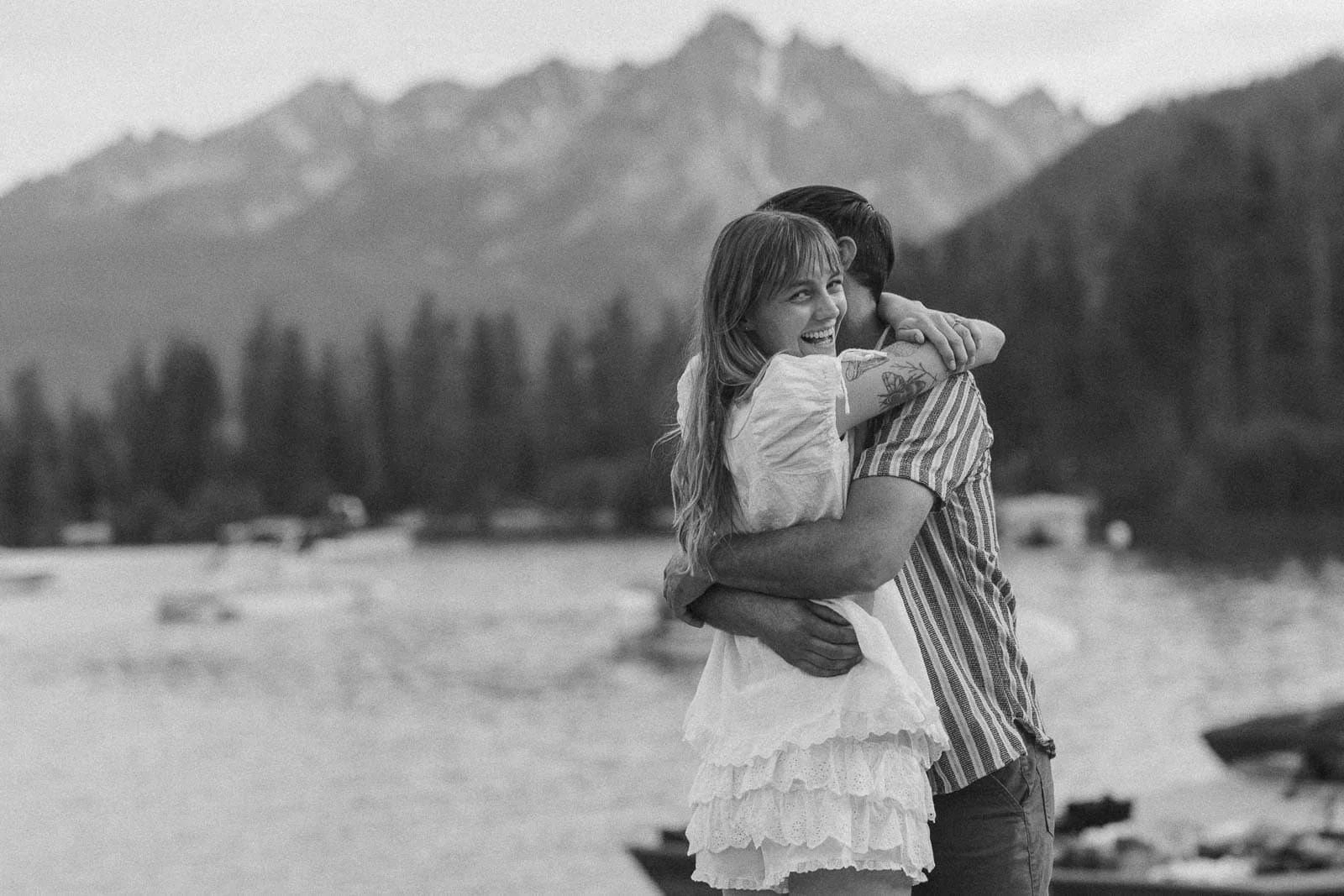  A woman in a white dress hugs a man by a lake, smiling at the camera. Mountains and trees are in the background. The photo is in black and white. 