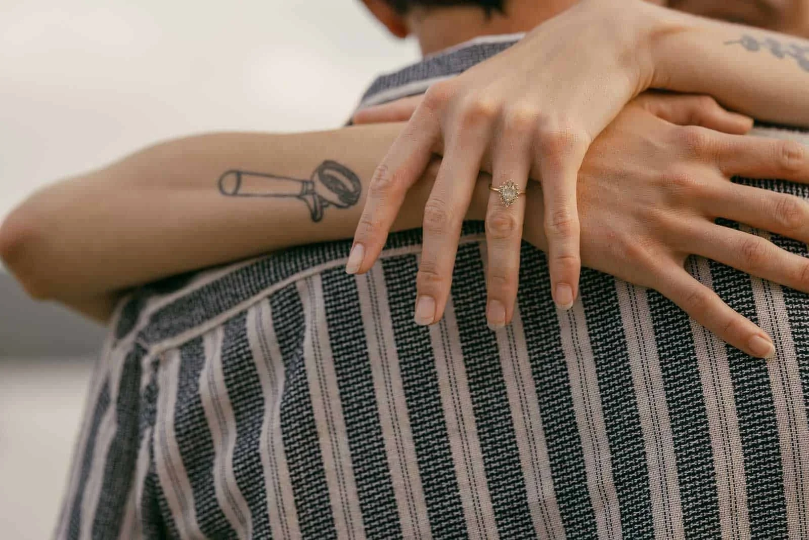  A close-up of a person hugging someone in a striped shirt, with their hand showing an engagement ring and a tattoo of a magnifying glass on their arm. 