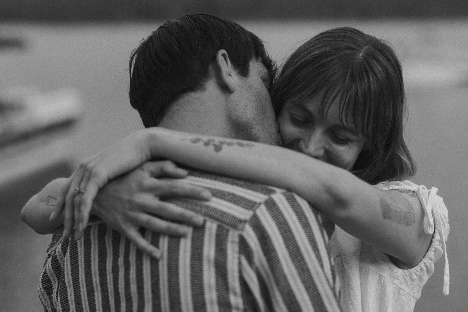  A black and white photo of a woman hugging a man closely, smiling with eyes closed. They are near water, and her arms are wrapped around his neck. 