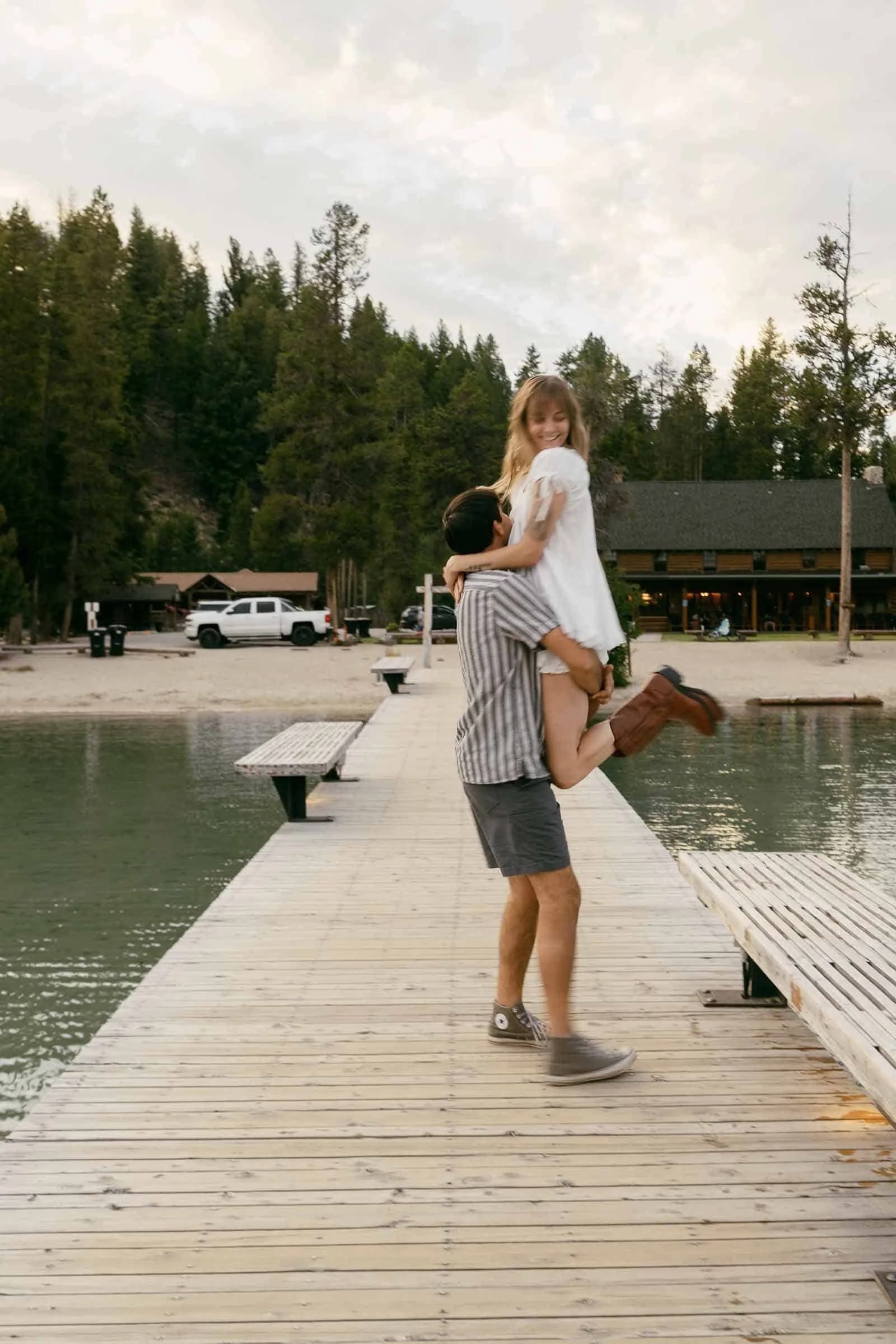  A man lifts a smiling woman in a white dress on a wooden dock by the water, with trees and a cabin in the background. 