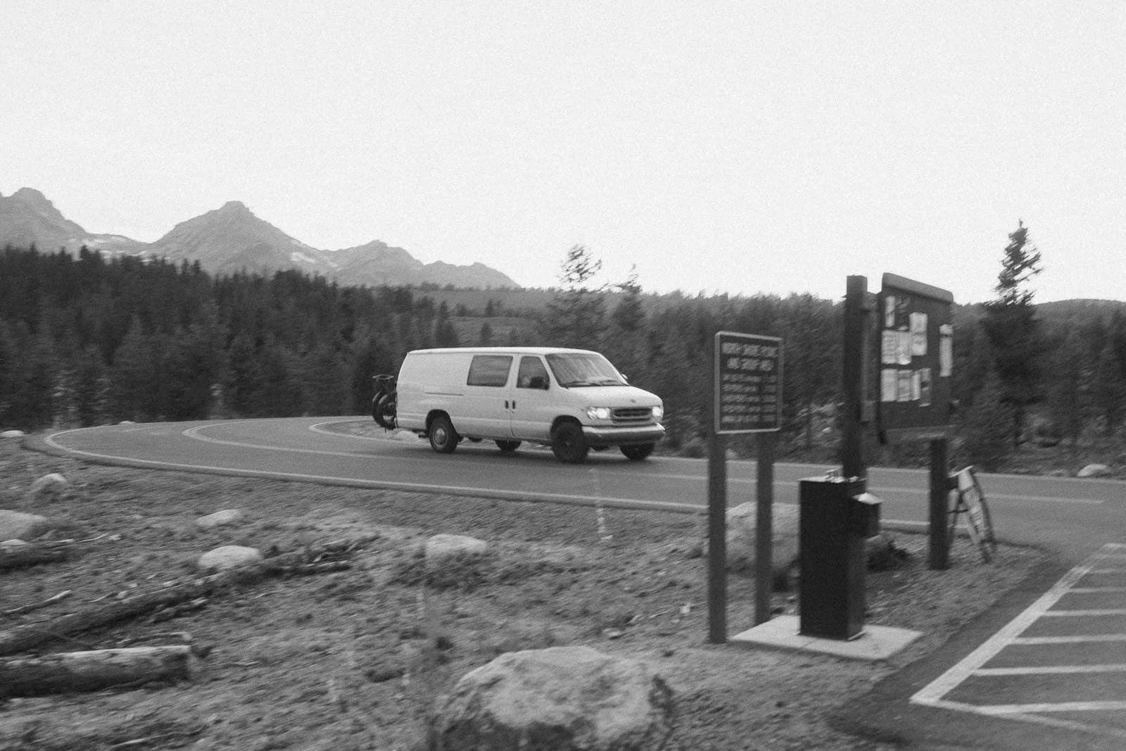  A white van drives on a curved road through a forested area with mountains in the background. A parking lot, information boards, and a bike are visible in the foreground. The image is in black and white. 