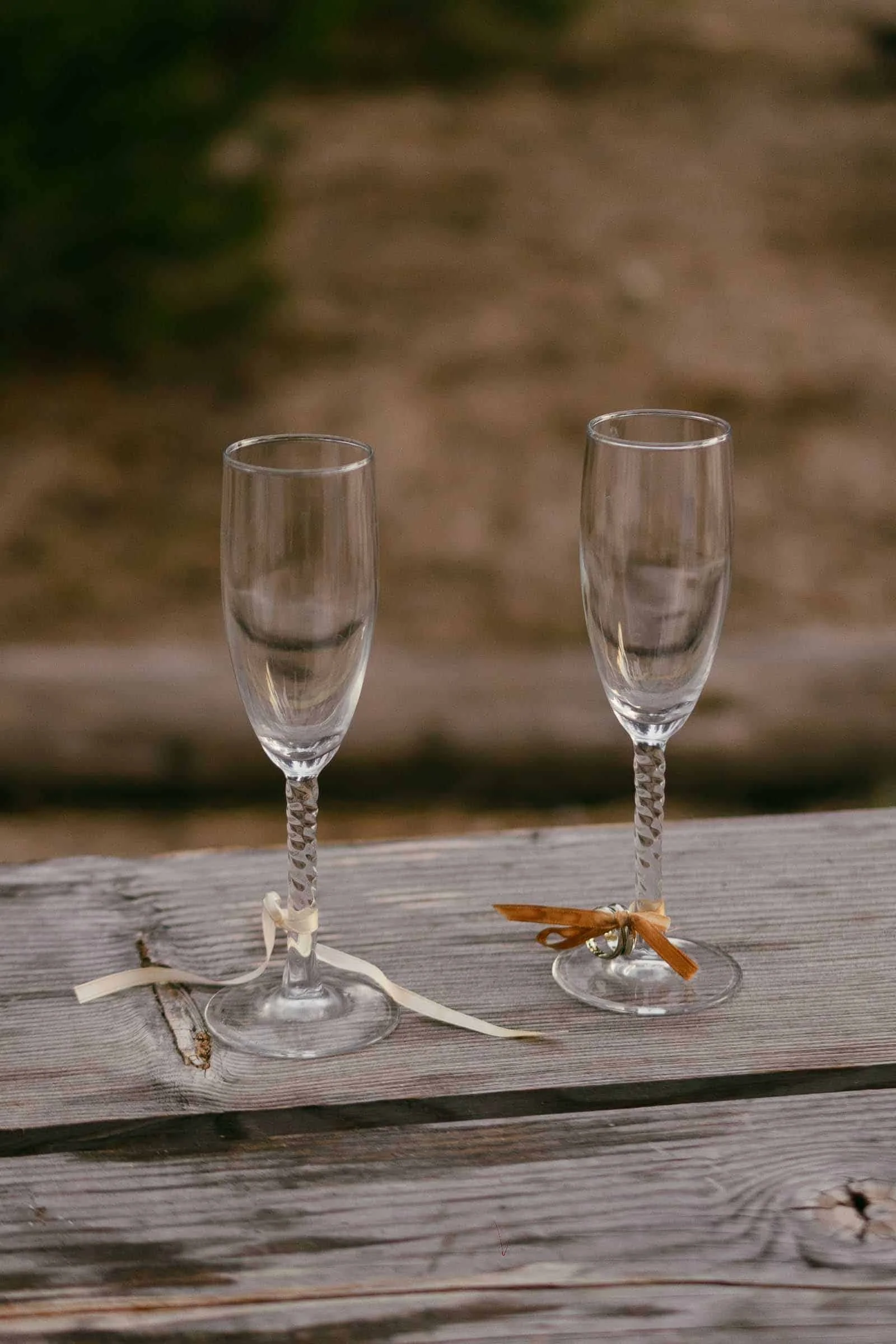  Two empty champagne flutes with decorative ribbons tied around the stems sit on a weathered wooden table outdoors. 
