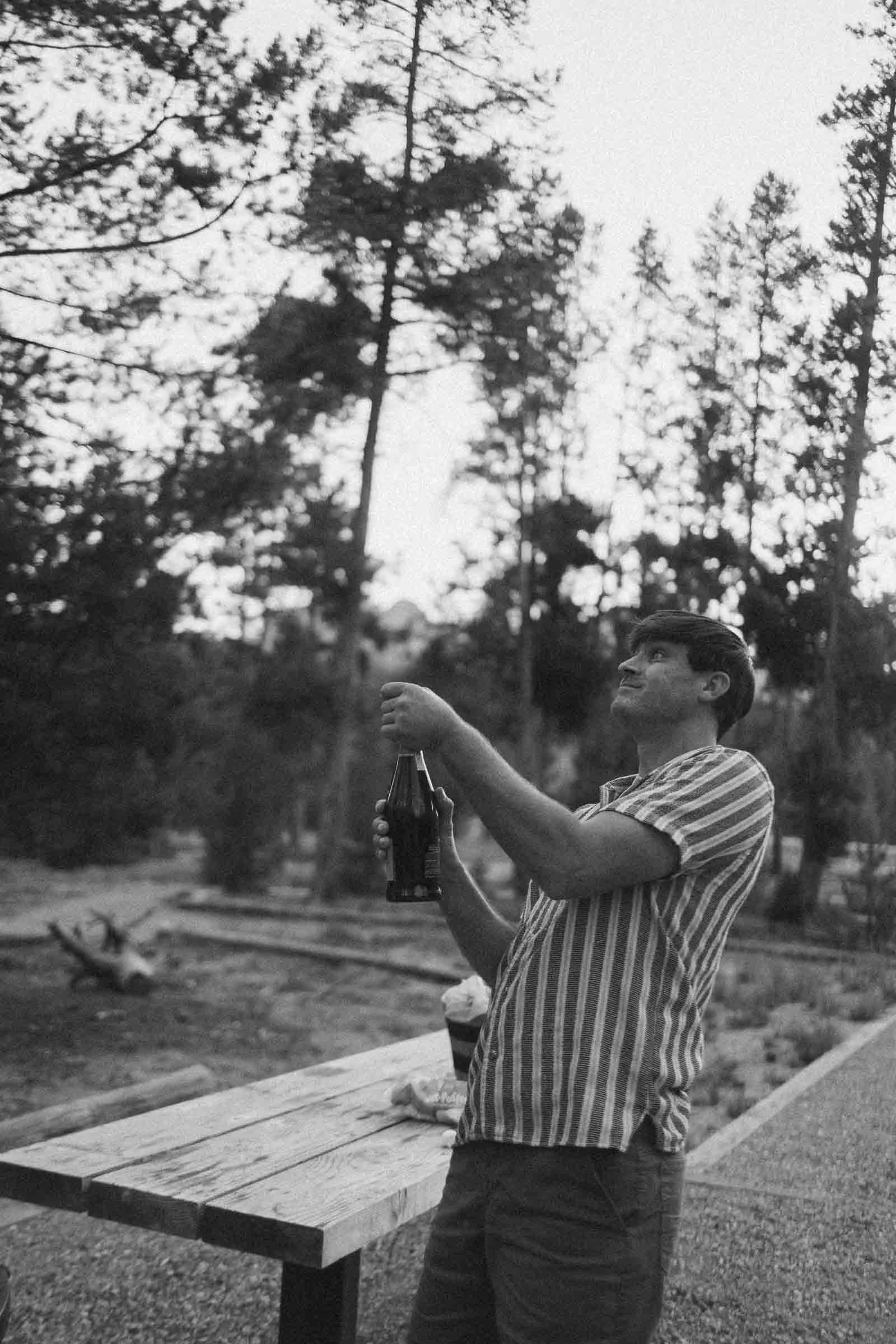  A person stands outdoors by a wooden table, opening a bottle and smiling. Trees fill the background, and the scene has a relaxed, casual atmosphere. The photo is in black and white. 
