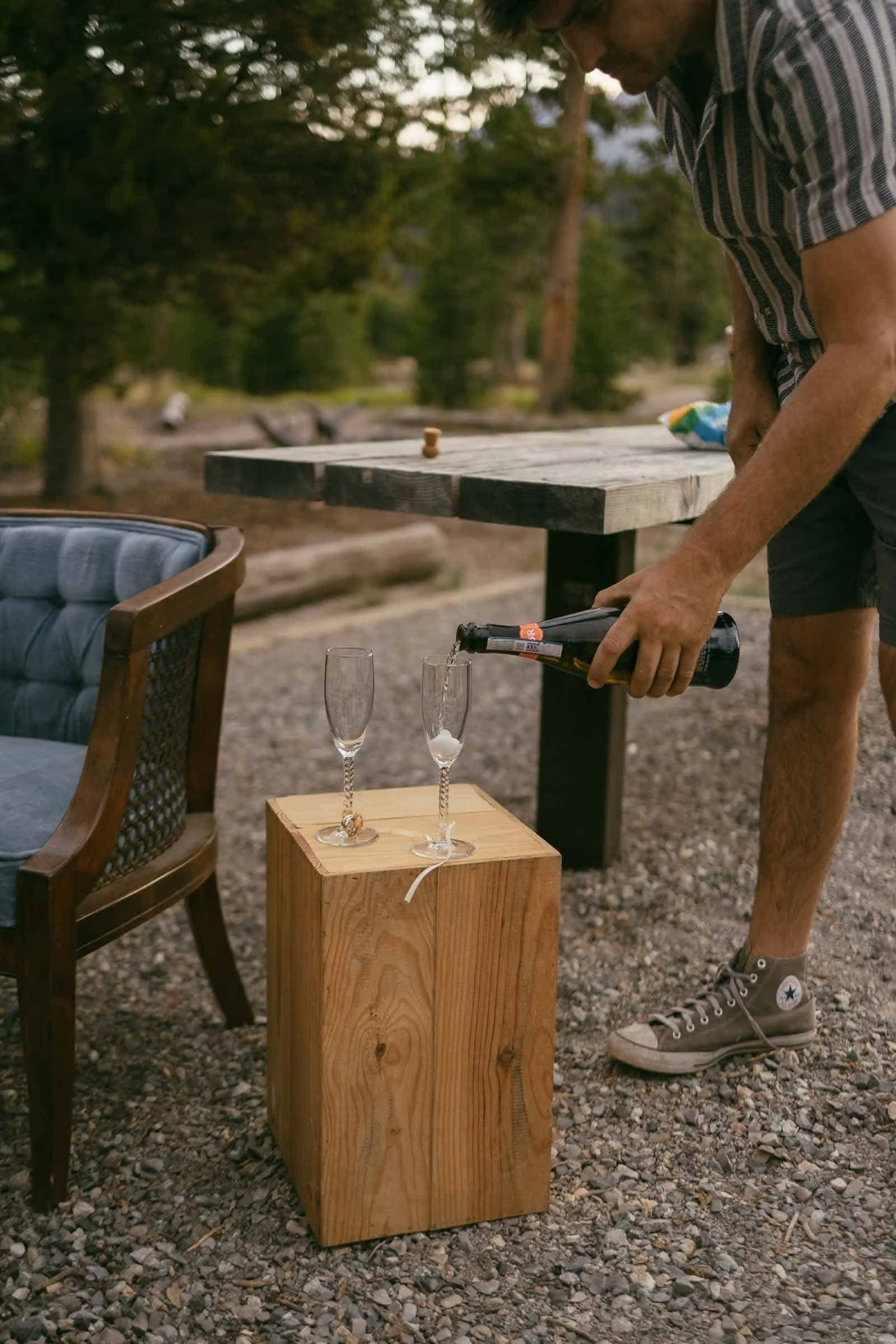  A person pours sparkling wine into two champagne flutes on a wooden block outside, with a chair and table nearby in a natural setting. 