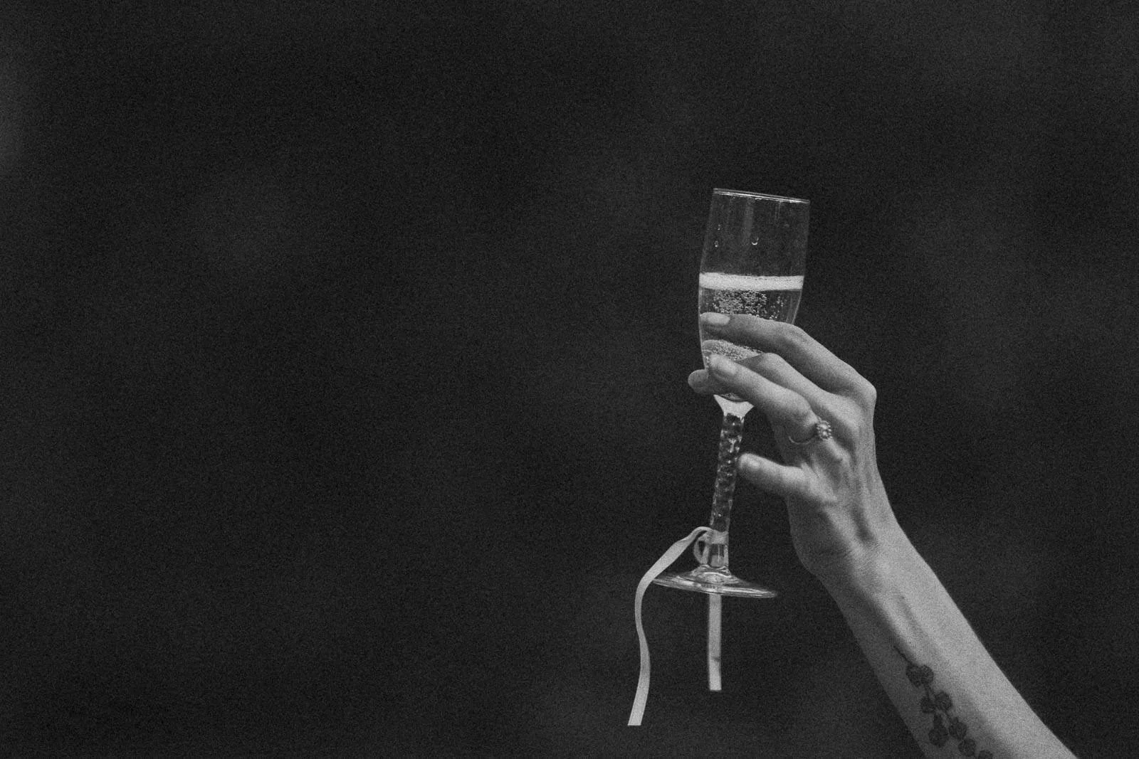 A tattooed hand raises a champagne flute with a ribbon tied around the stem, set against a dark, softly blurred background.
