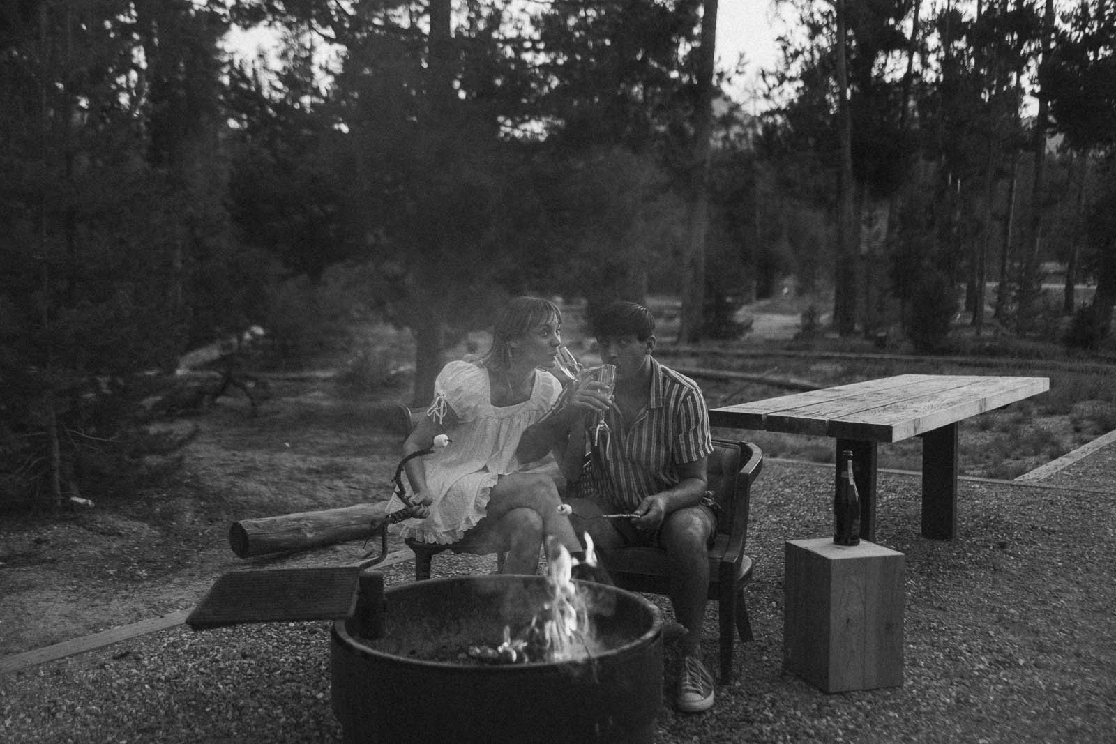  A couple sits by a campfire roasting marshmallows. They smile and lean in close, surrounded by trees, with a picnic table and bottle nearby. The image is in black and white. 