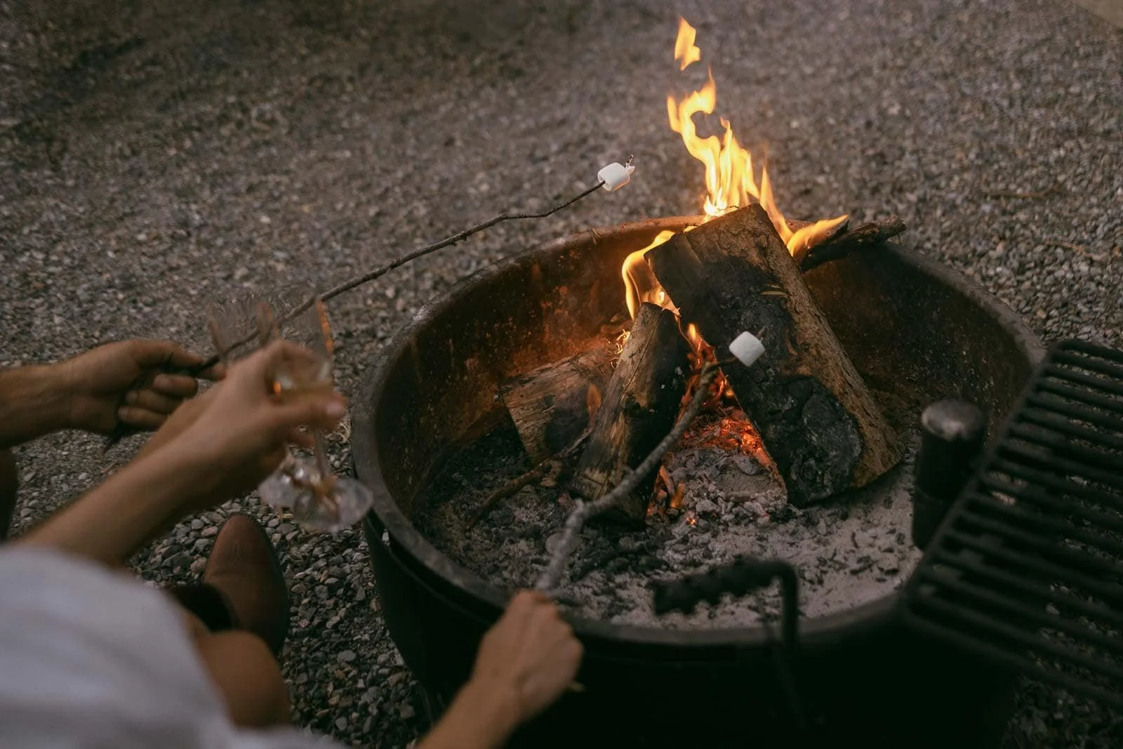  Two people roast marshmallows over a campfire in a fire pit. One person holds a glass, while both use sticks to toast the marshmallows above the flames. Gravel surrounds the fire pit. 
