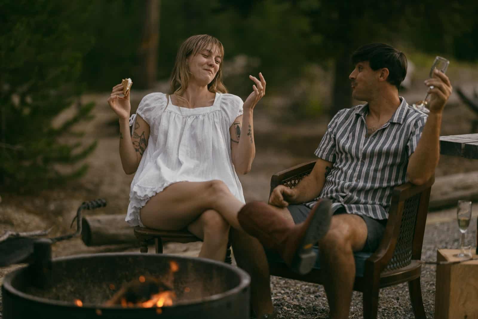  Two people sit by a campfire outdoors. One, in a white dress, is holding a s’more and smiling with their eyes closed. The other, in a striped shirt, looks at them, holding a drink. Both appear relaxed and happy. 