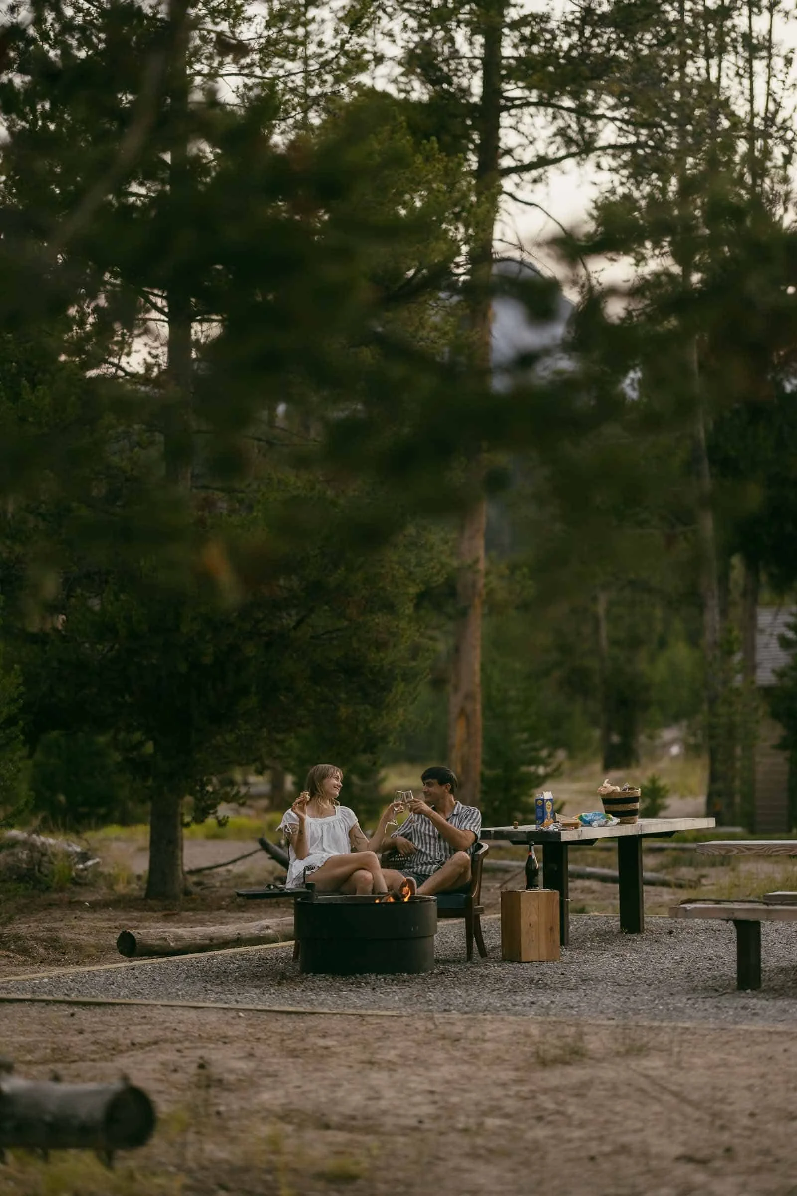  A couple sits by a campfire in a wooded area, talking and relaxing. There is a picnic table nearby with food and drinks, surrounded by tall trees and soft evening light. 