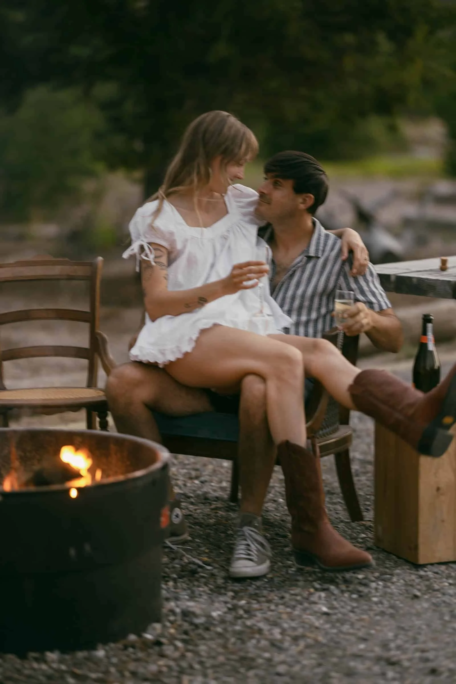  A woman in a white dress and brown boots sits on a man’s lap, both smiling at each other by a campfire. They hold drinks, with a wine bottle on a small table nearby. The scene is outdoors with blurred greenery. 