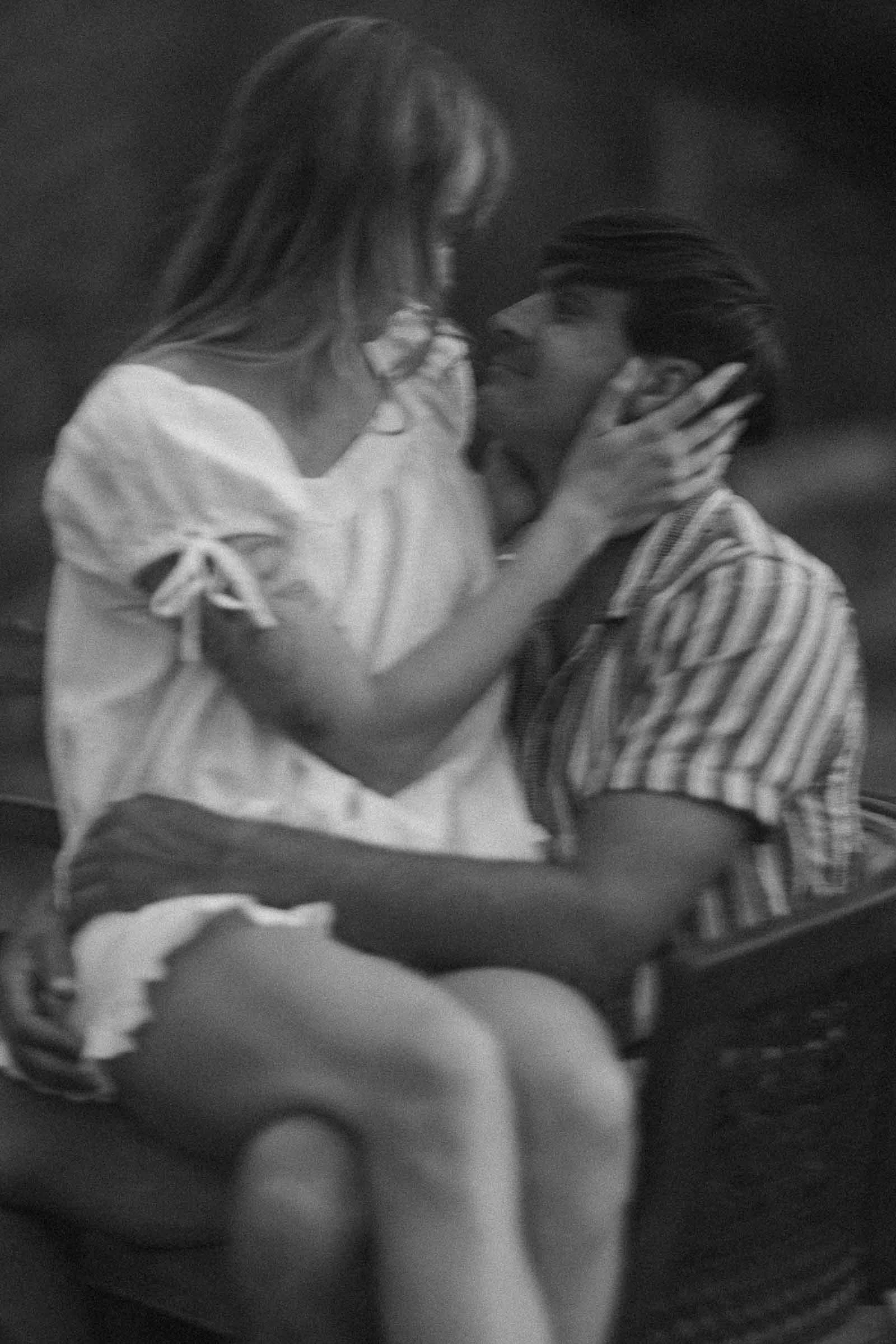  A black and white, blurred photo of a couple sitting close together. The woman sits on the man’s lap, facing him, as they hold each other’s faces and gaze into each other’s eyes affectionately. 