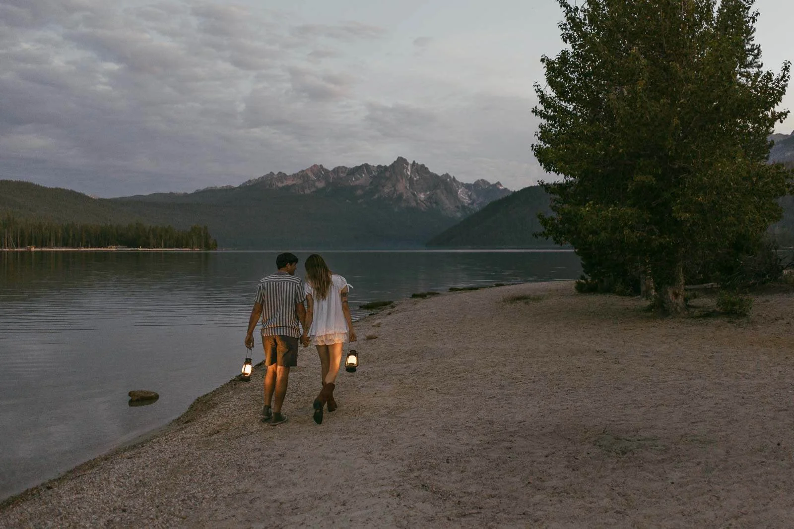 At dusk, a couple carries lanterns along the sandy shore of a calm lake, the mountains and trees behind them mirrored in the still water.
