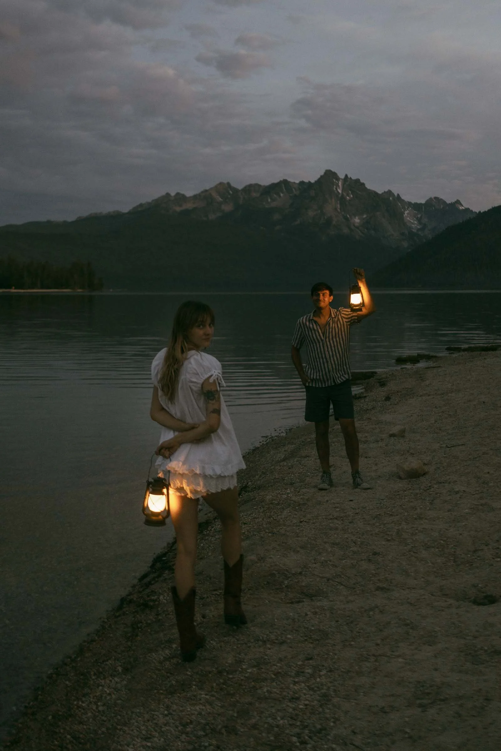  A woman in boots holds a lantern on a lakeshore at dusk, looking back toward a man holding another lantern. Mountains and still water are in the background under a cloudy sky. 