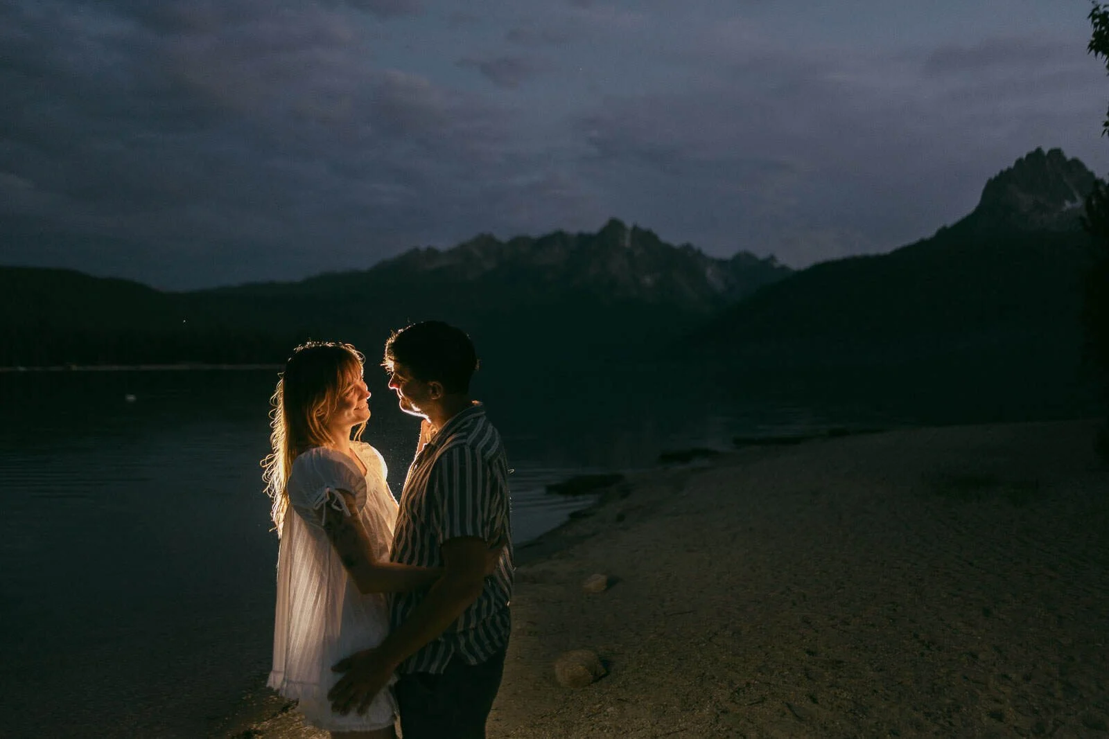  A couple stands on a sandy lakeshore at dusk, facing each other and smiling, with gentle light illuminating them. Mountains and a calm lake are in the background under a cloudy sky. 