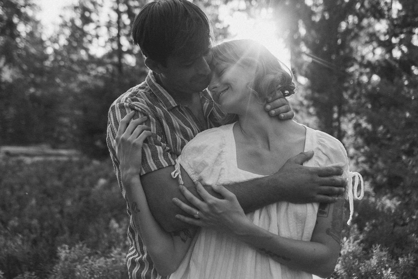 In black and white, a woman in a light dress and a man in a striped shirt embrace among sunlit trees, laughing as they look at each other.