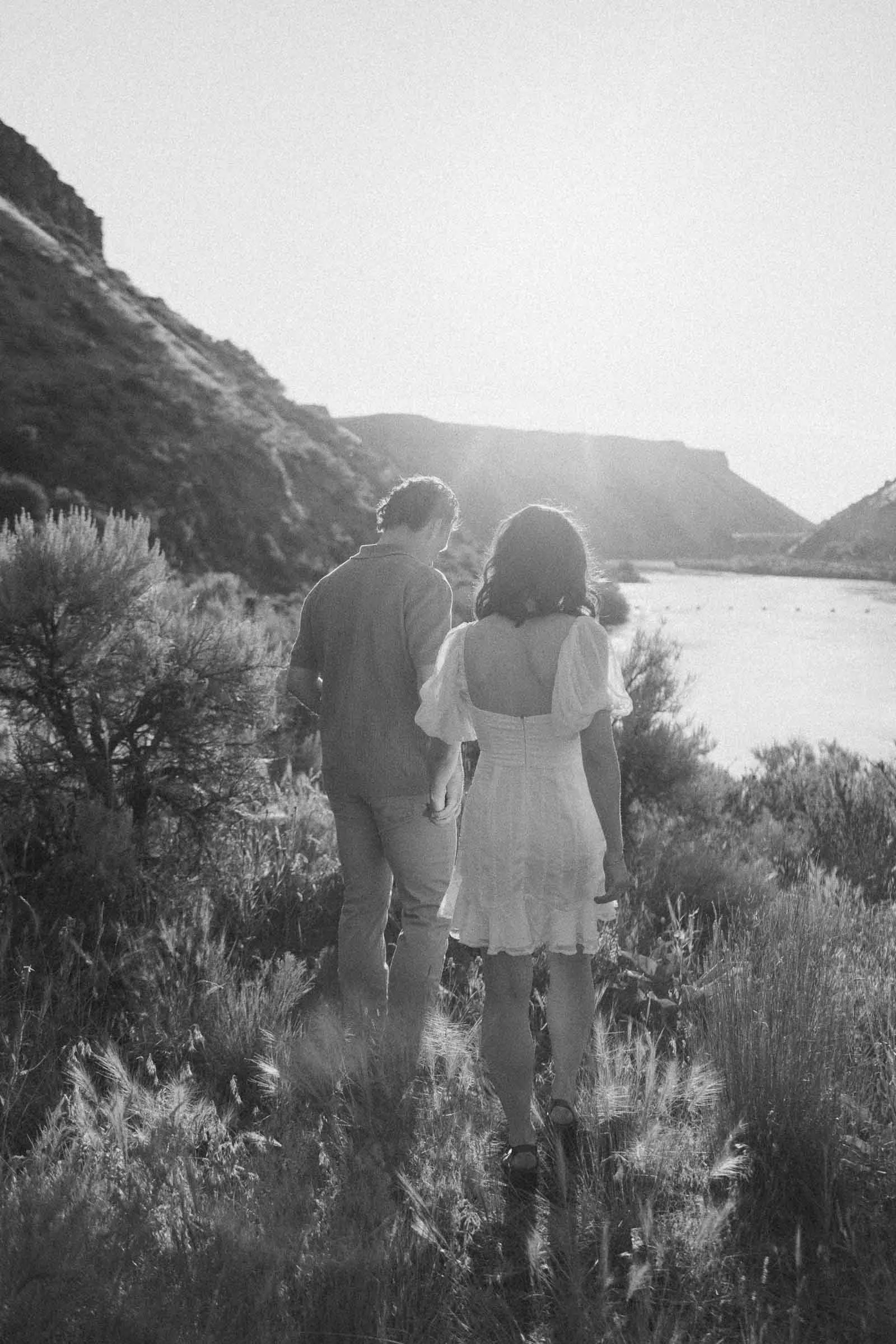  A man and woman holding hands walk together along a grassy trail near a river. 