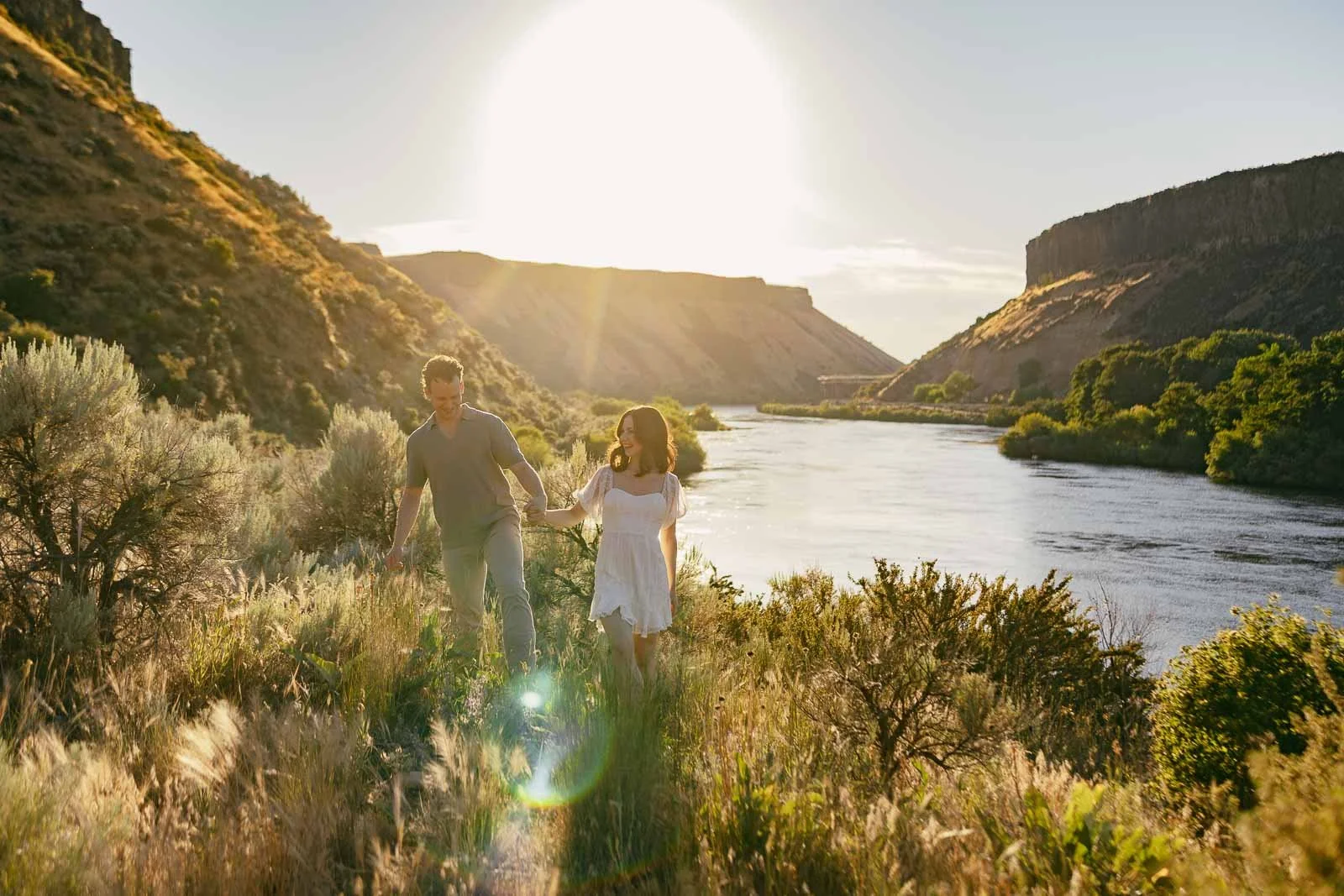 An engaged couple walks hand in hand through sagebrush and tall grass beside the Boise River at golden hour during a Lucky Peak engagement session.