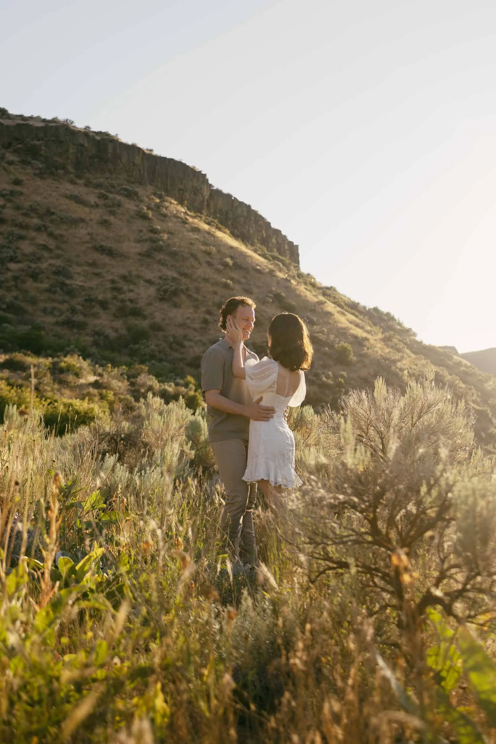  A couple embraces in a sunlit, grassy field beneath a hillside. 