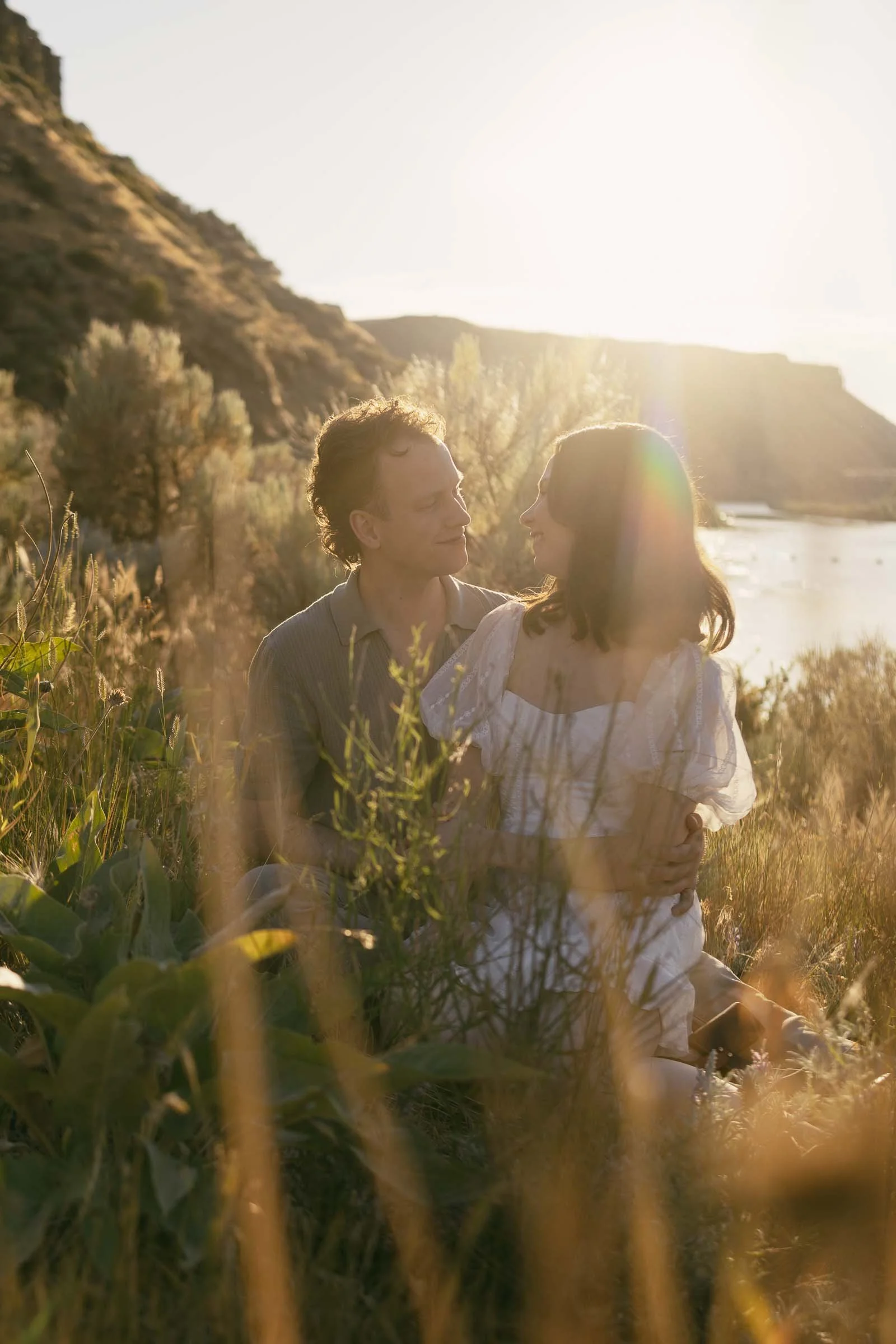  A couple sits closely and smiles at each other in a grassy field during sunset. 