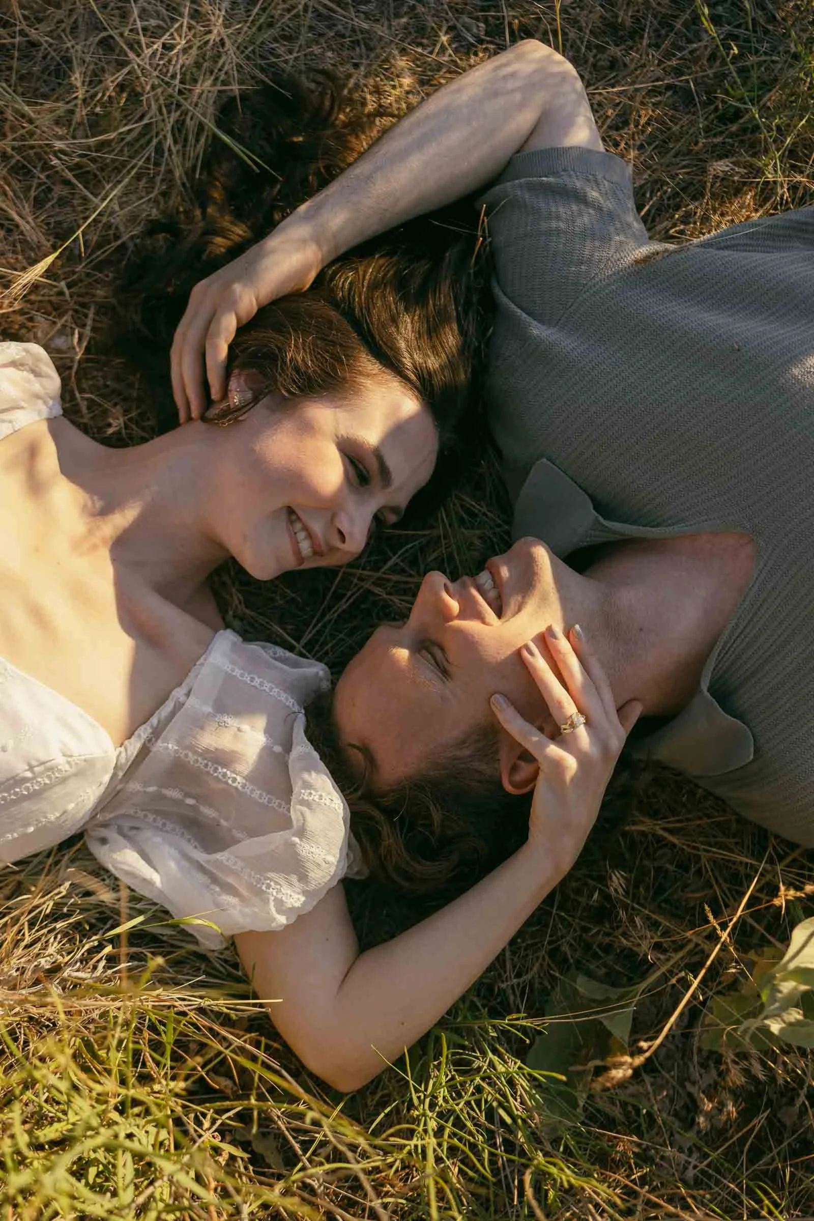  A couple lying in a field of dry grass, smiling at each other while touching faces. 