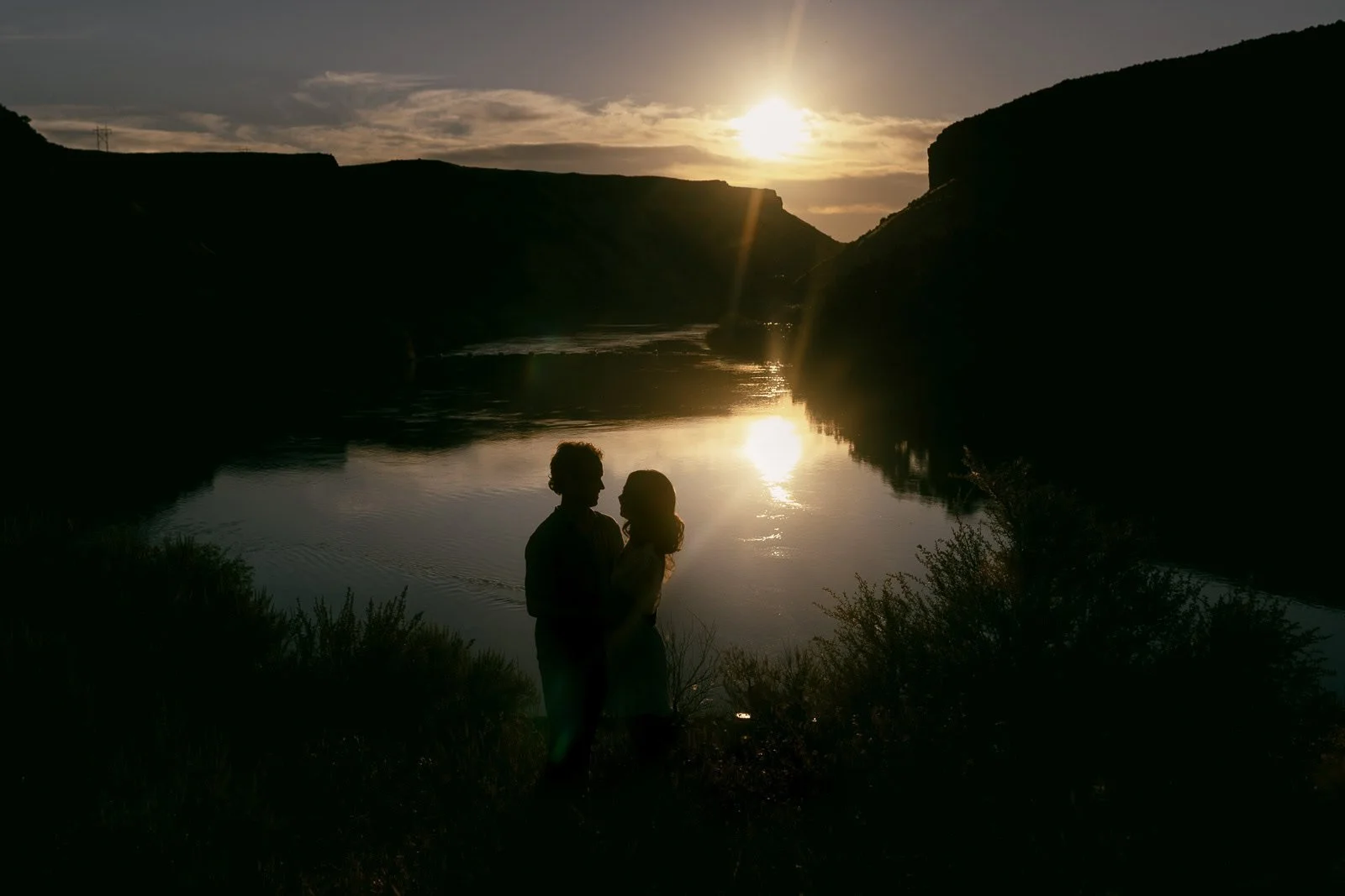  A couple stands in silhouette against the sunset over a river flowing between canyon walls. 
