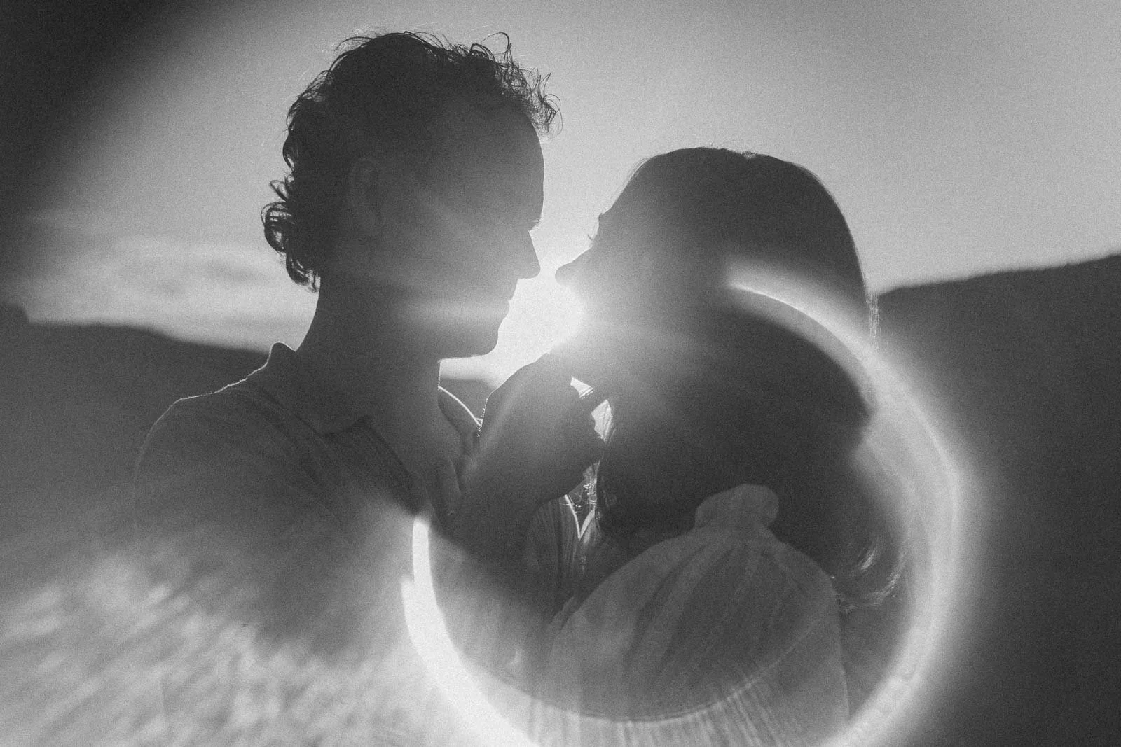  A couple silhouetted against a bright sun in a black and white film-style photograph. 