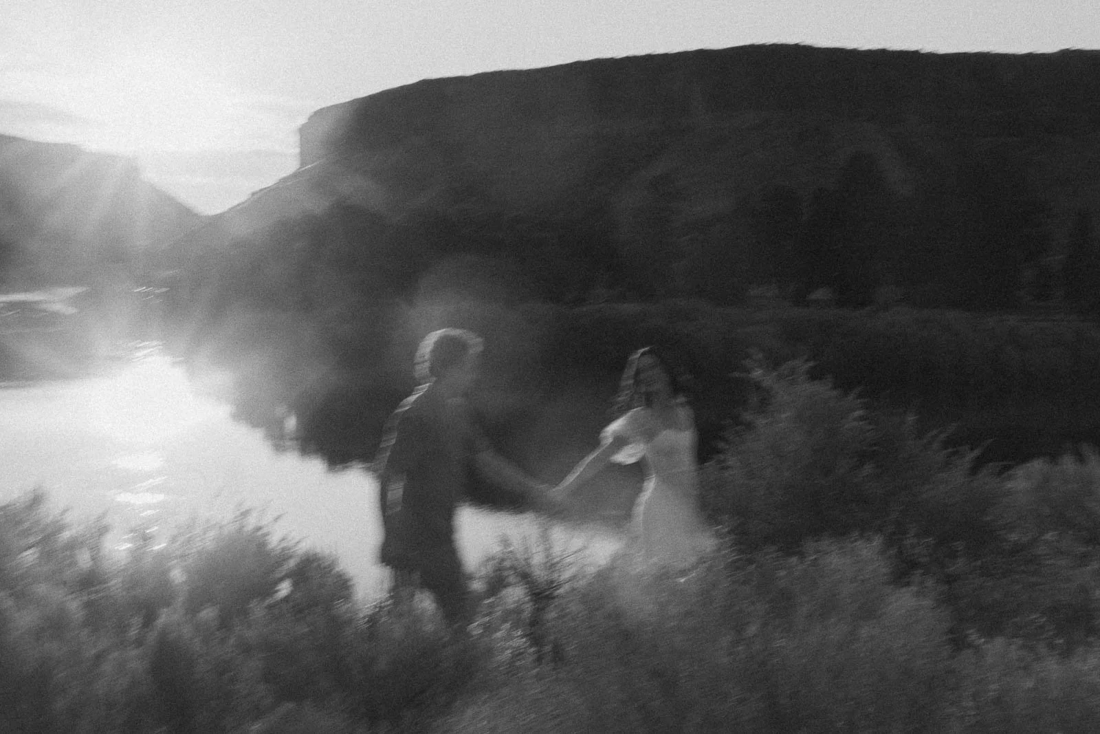  A couple holding hands walks through tall grass by a river at sunset. 