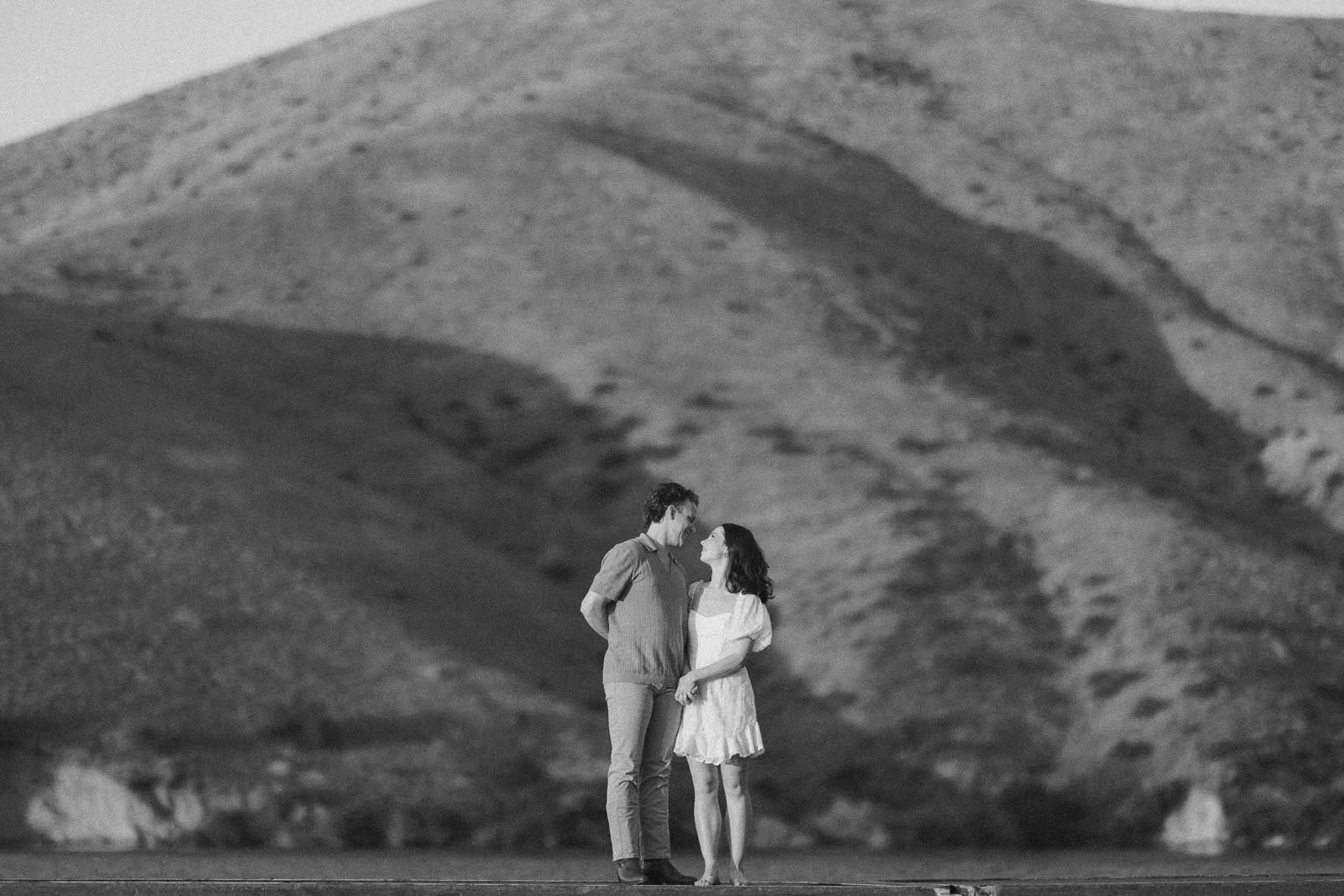  A man and a woman in light clothing standing face-to-face and holding hands in front of a large, barren hill. 