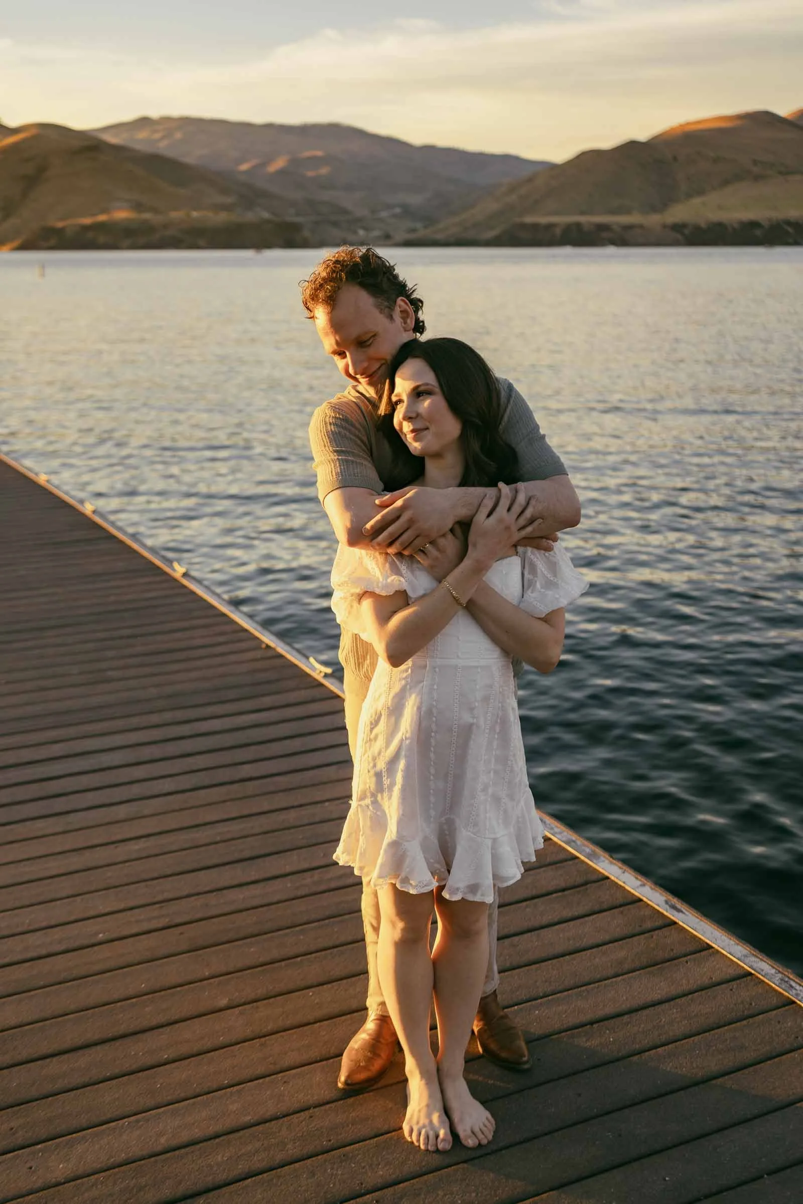  A couple embraces on a wooden dock overlooking a lake at sunset. 