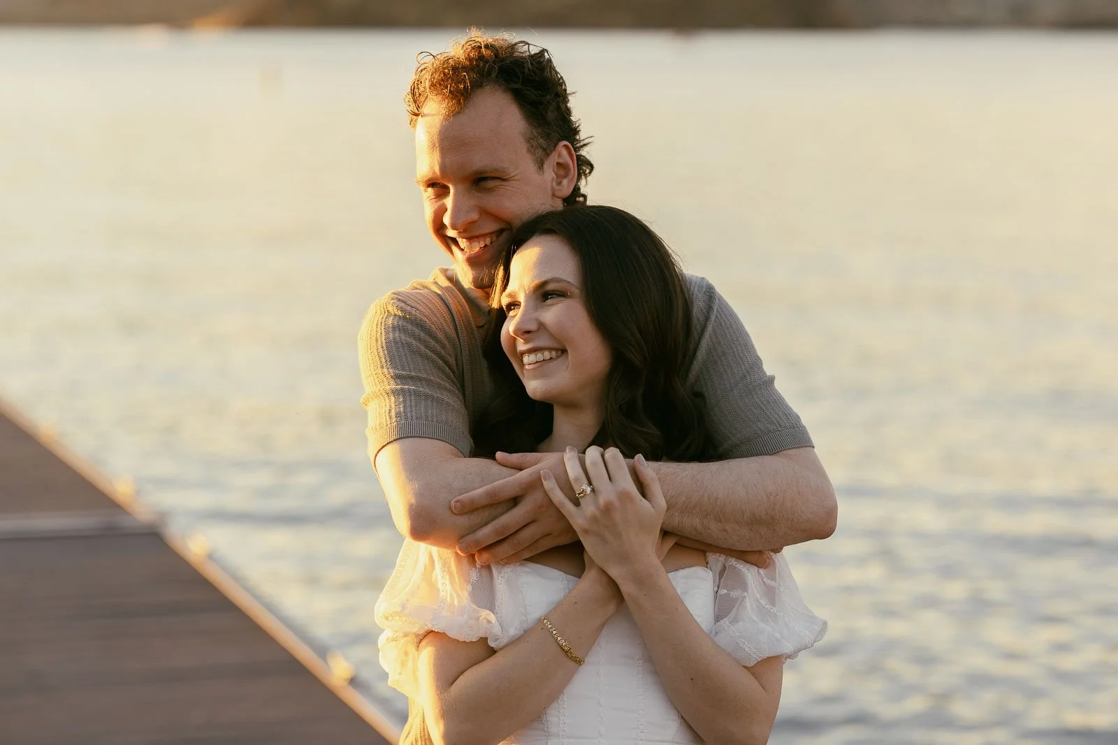 A man embraces a woman from behind as they both smile while standing on a pier at sunset. 