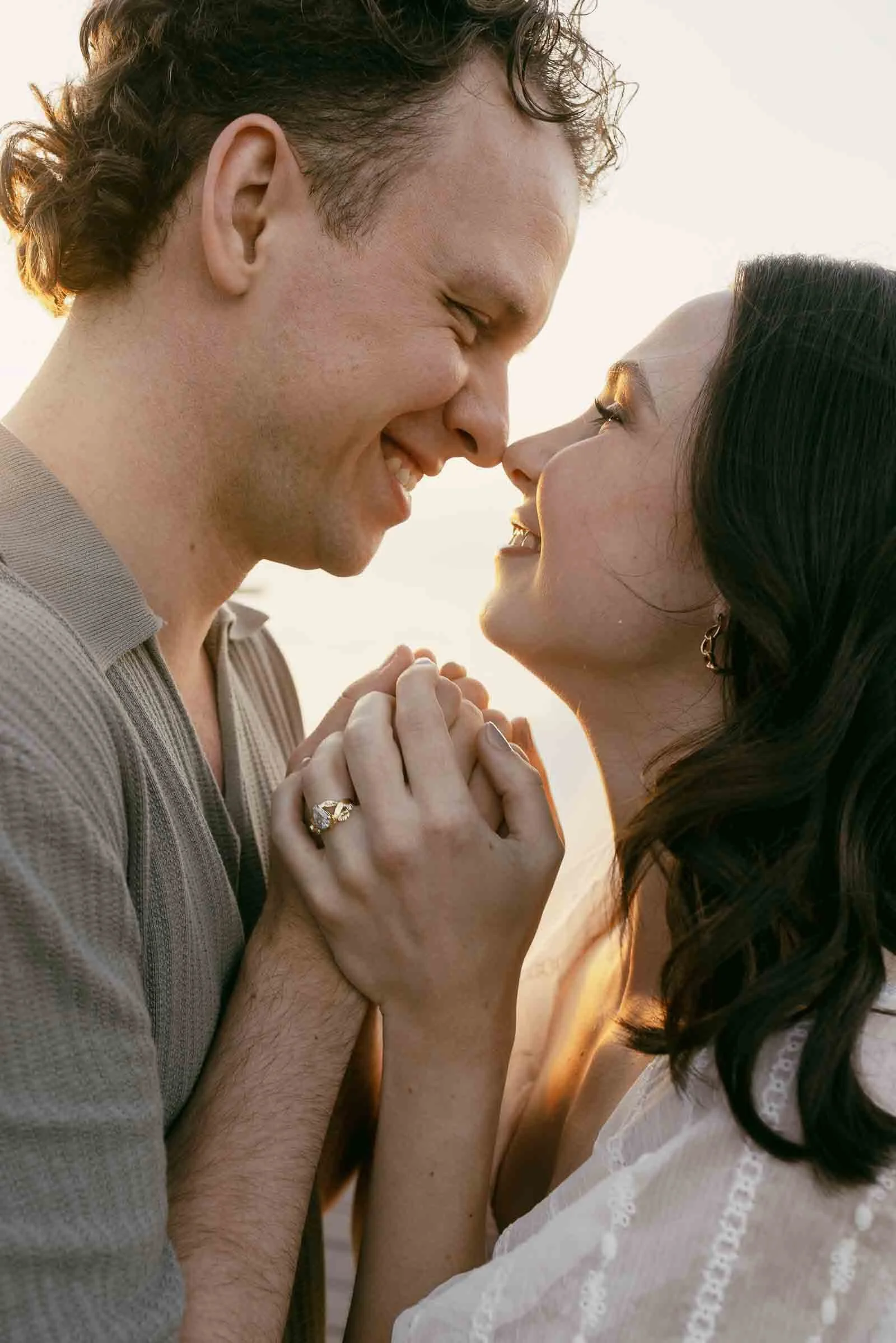  A couple smiling at each other while holding hands, with a prominent diamond engagement ring visible on the woman’s finger. 