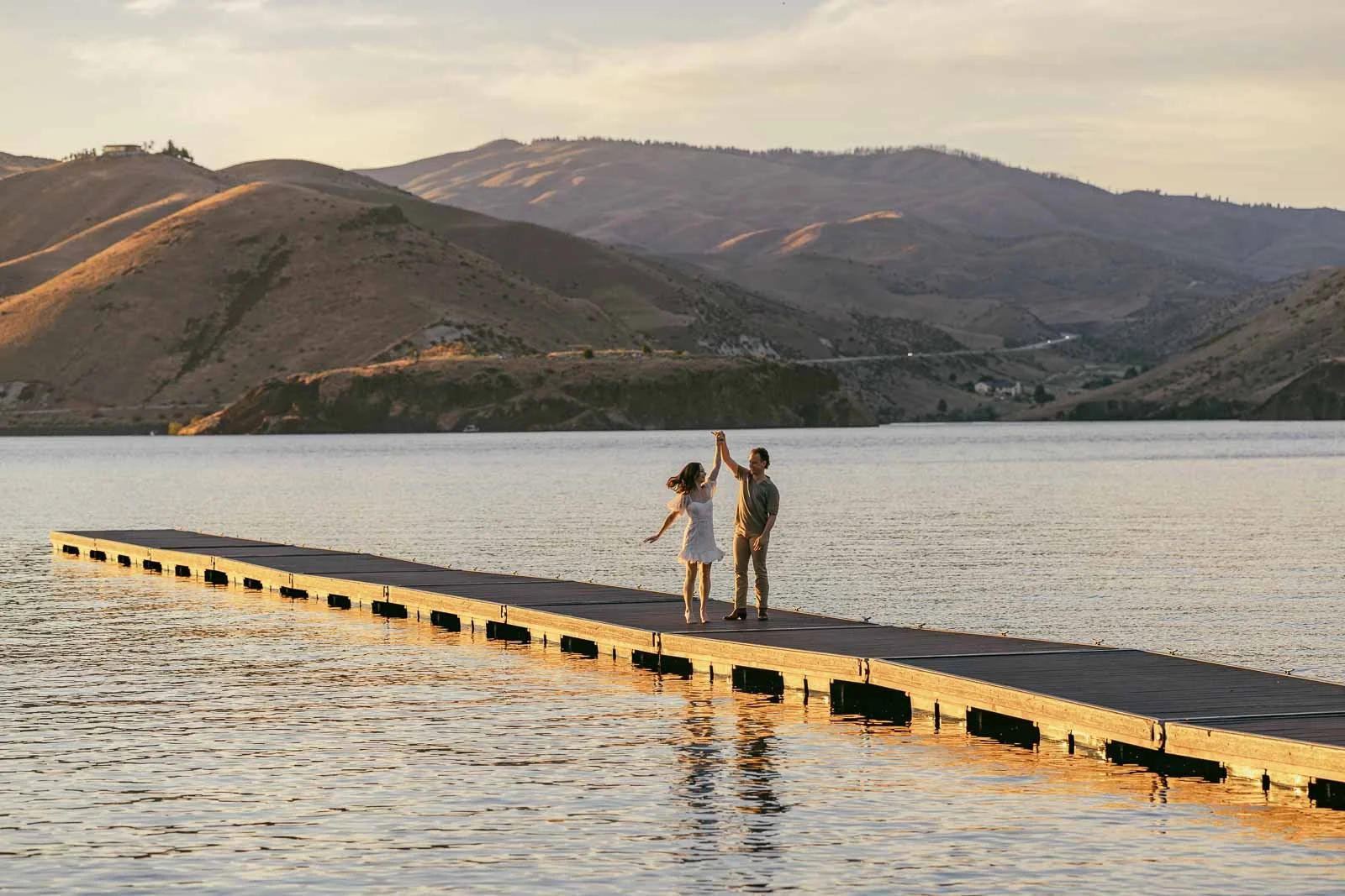 A man twirls his fiancée on a long floating dock on Lucky Peak Reservoir as sunset lights the Boise foothills behind them in a dance-inspired engagement photo.