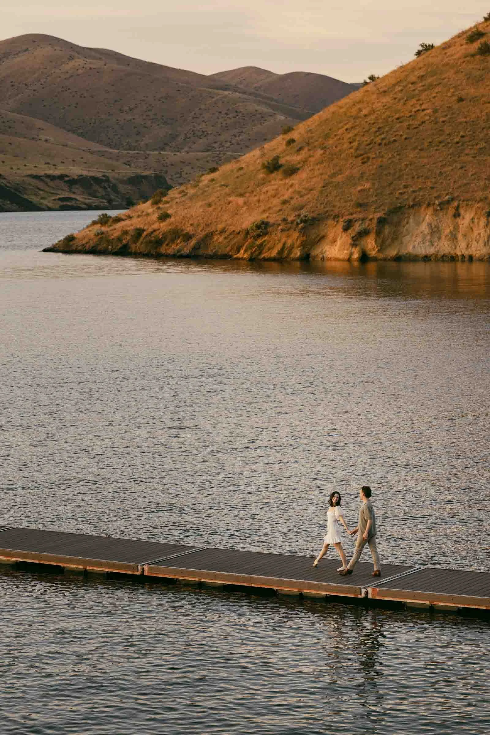  A couple holding hands walks along a floating dock on a lake with hills in the background. 