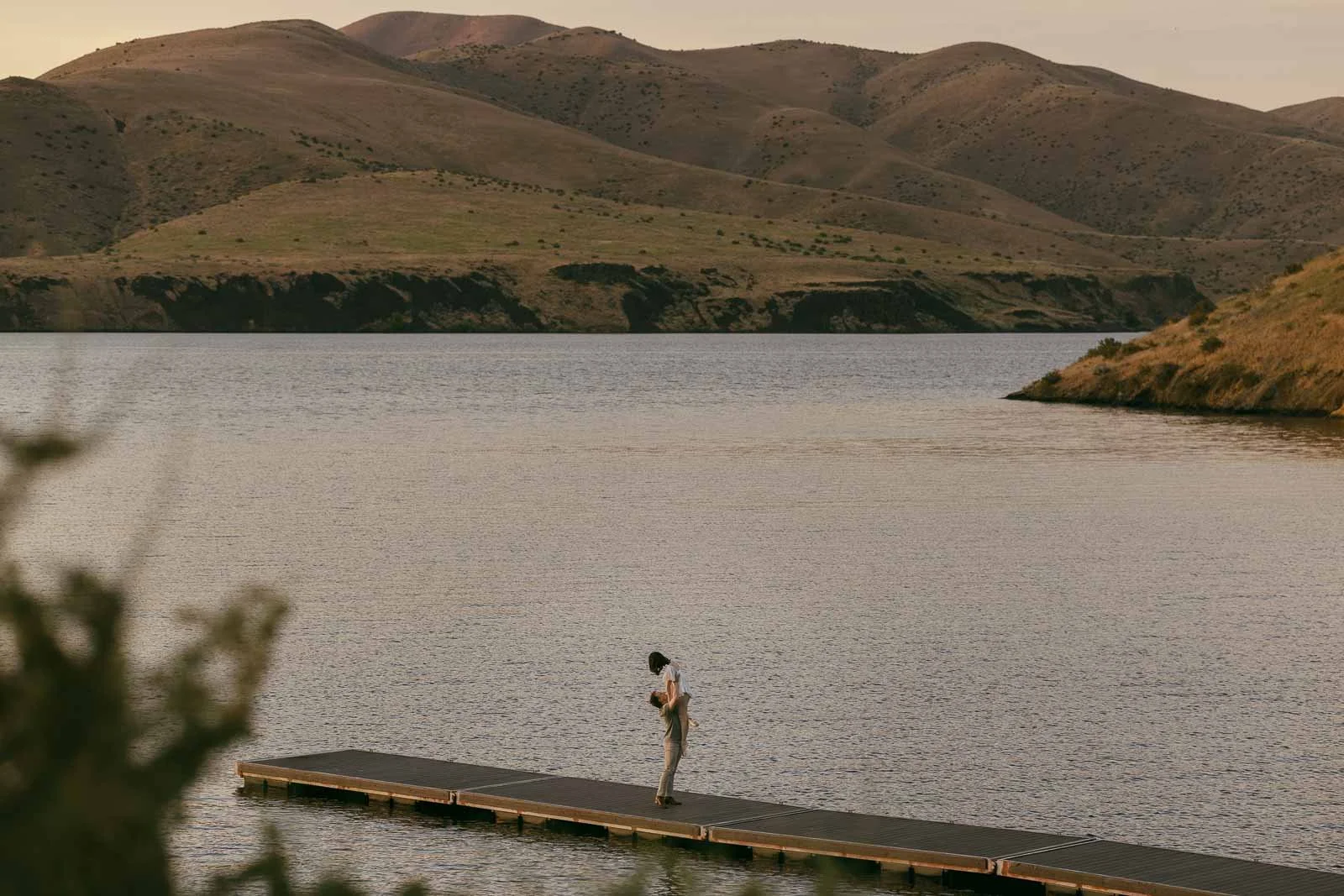 A couple embraces on a long wooden dock extending into a lake surrounded by hills. 