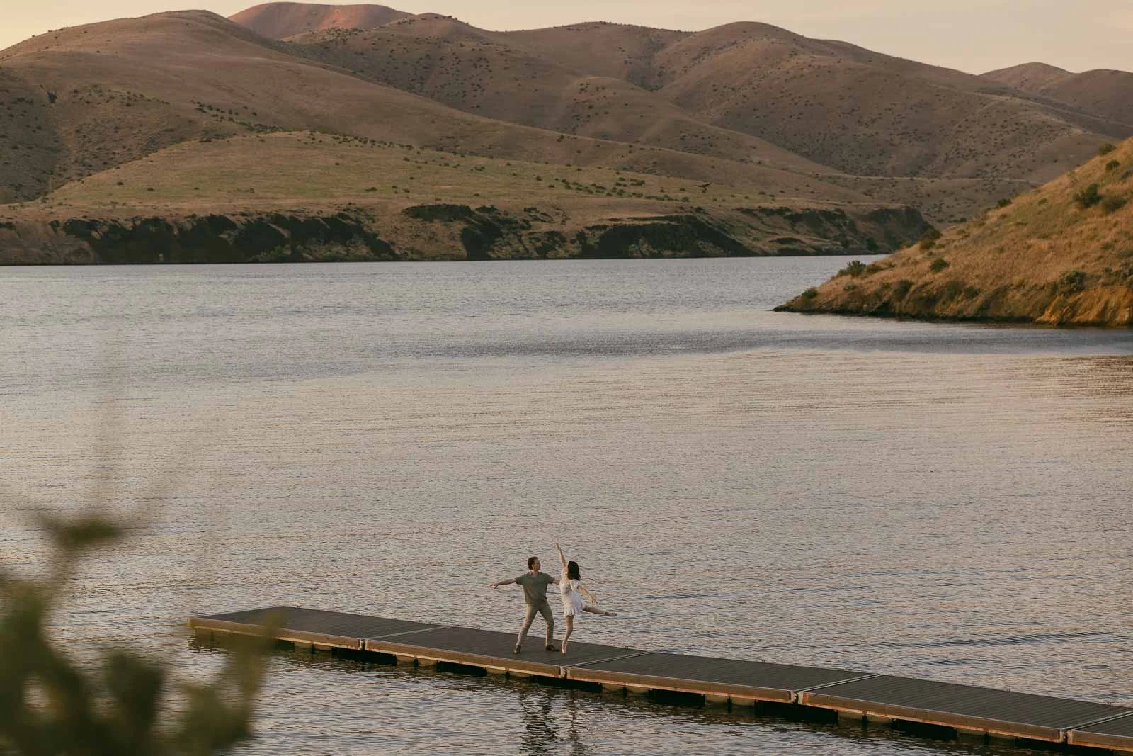  A couple dances on a floating dock in the middle of a lake surrounded by hills. 