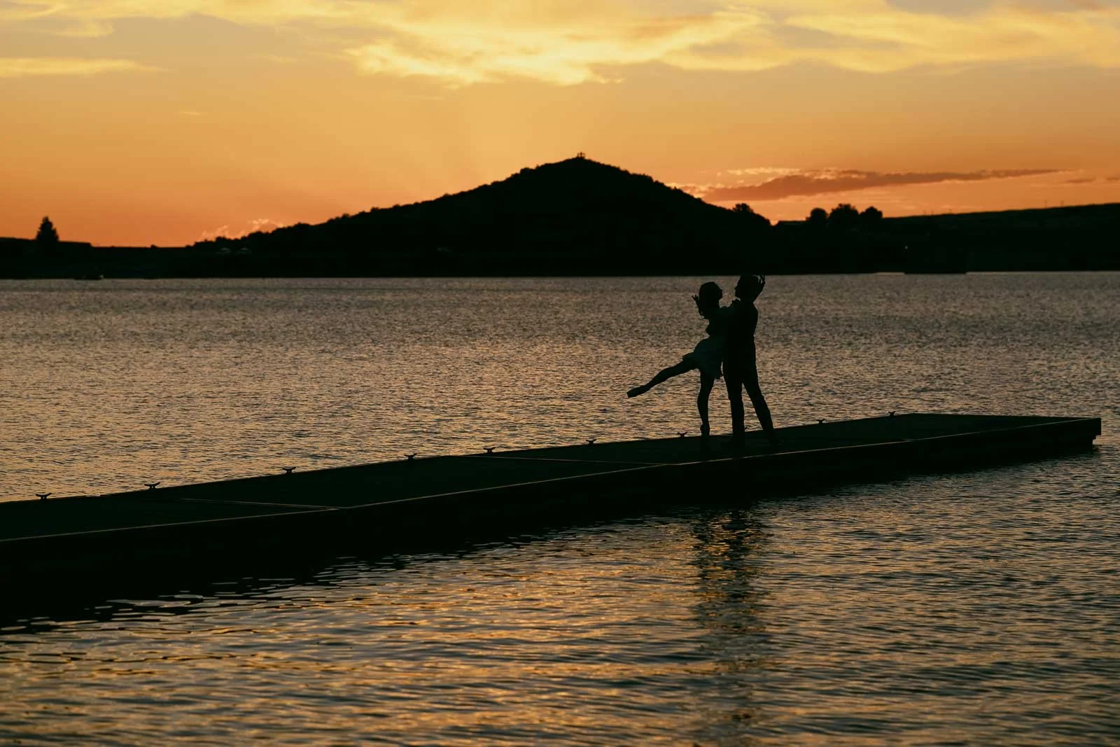  Silhouettes of a couple dancing on a pier at sunset over a lake. 
