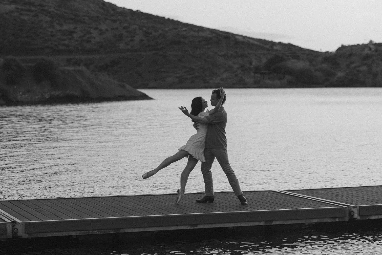 Black and white photo of a ballerina en pointe, extending one leg behind her as her fiancé, in cowboy boots, holds her close on a dock at Lucky Peak.