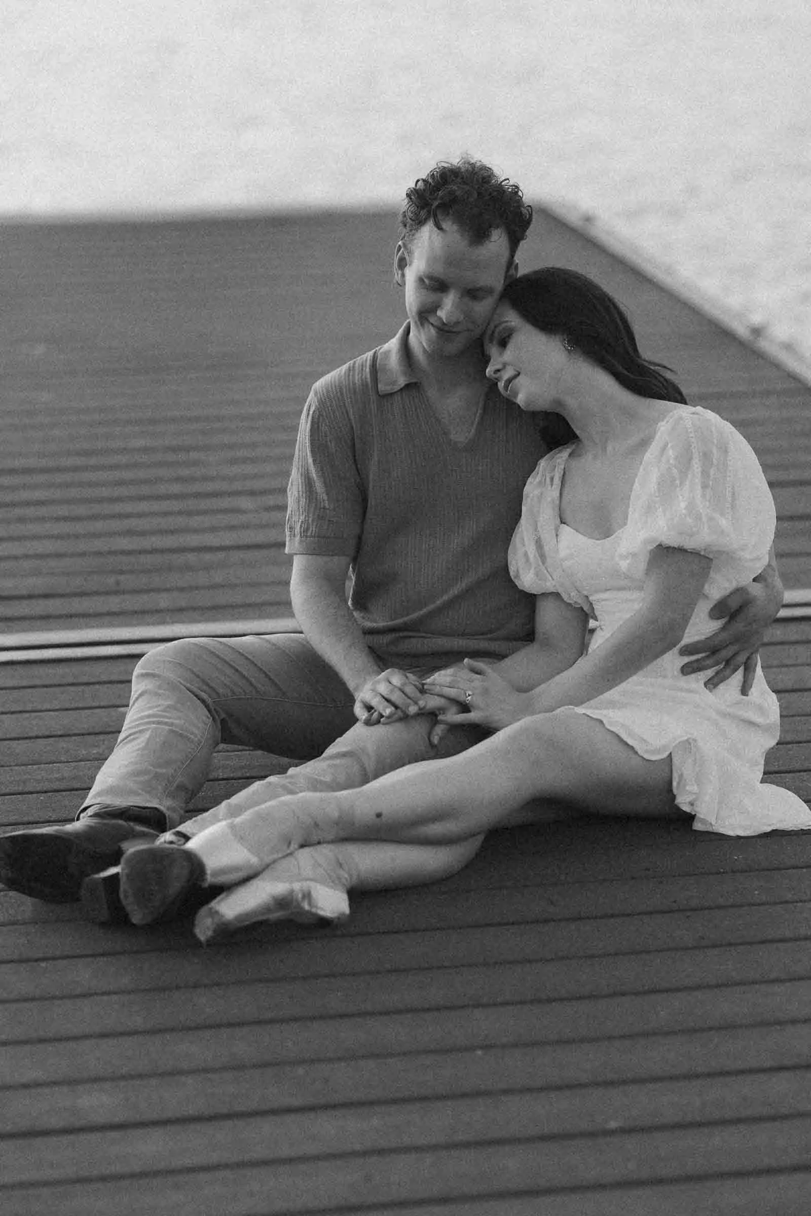  A couple sits together on a wooden dock, the woman leaning her head on the man’s shoulder while holding his hand. 