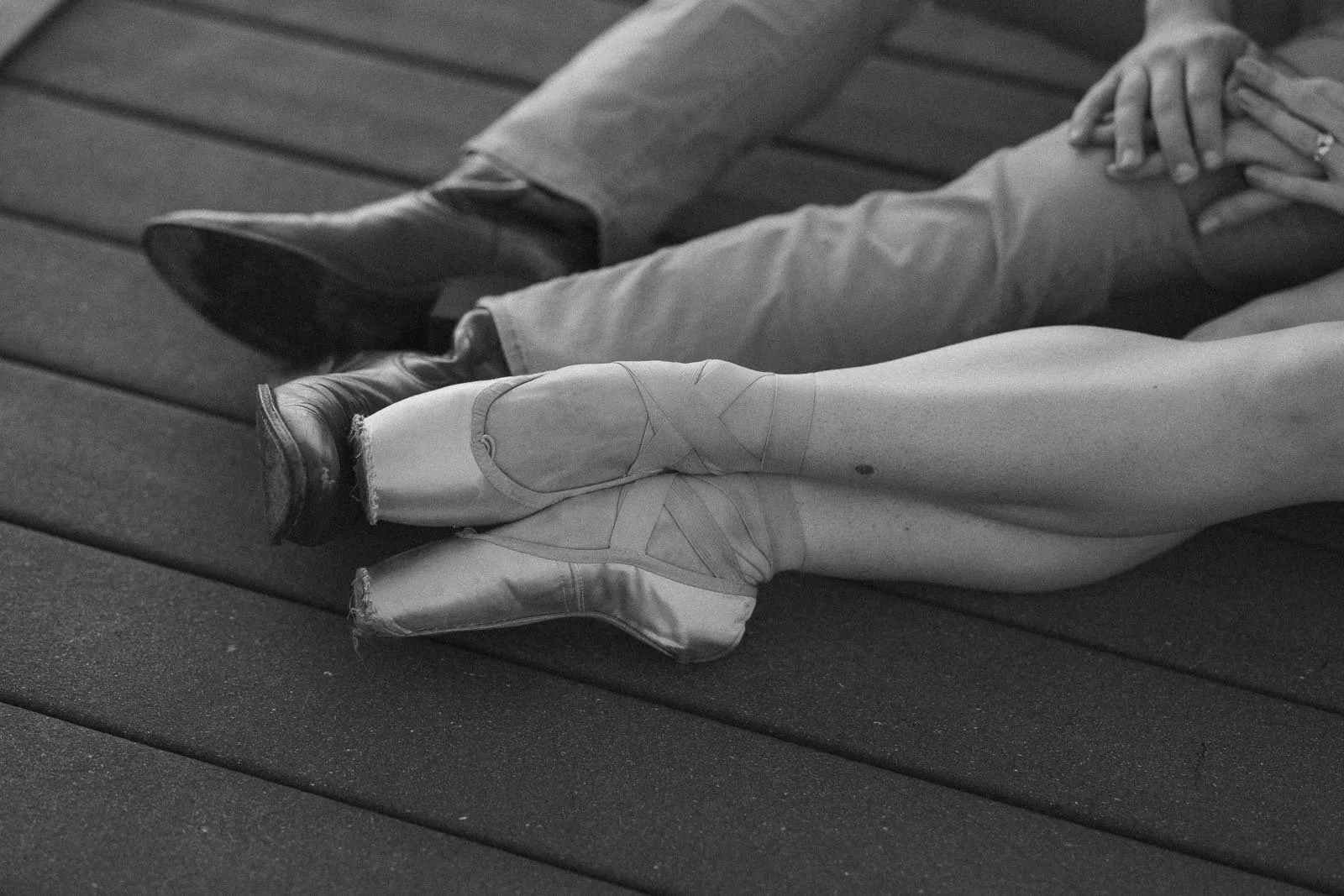  A pair of feet in ballet slippers resting near a pair of feet in cowboy boots, with hands held together above. 