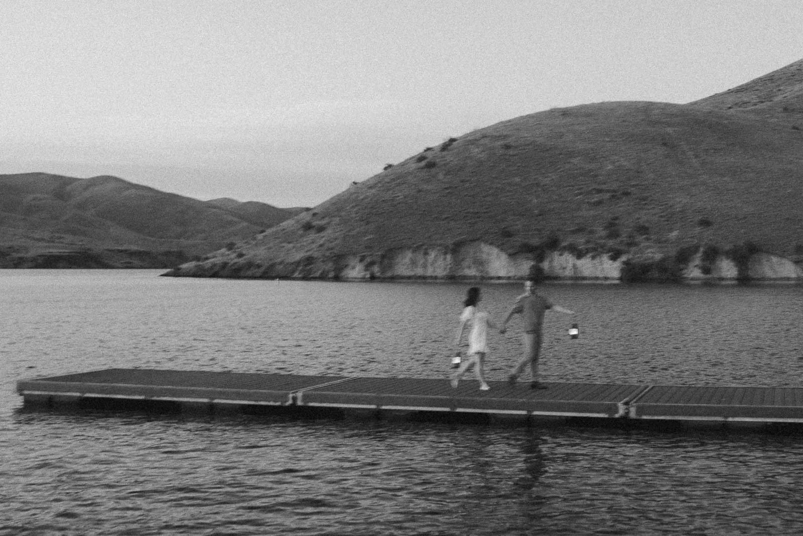  A couple holding hands and carrying lanterns walk across a floating dock on a lake surrounded by hills. 