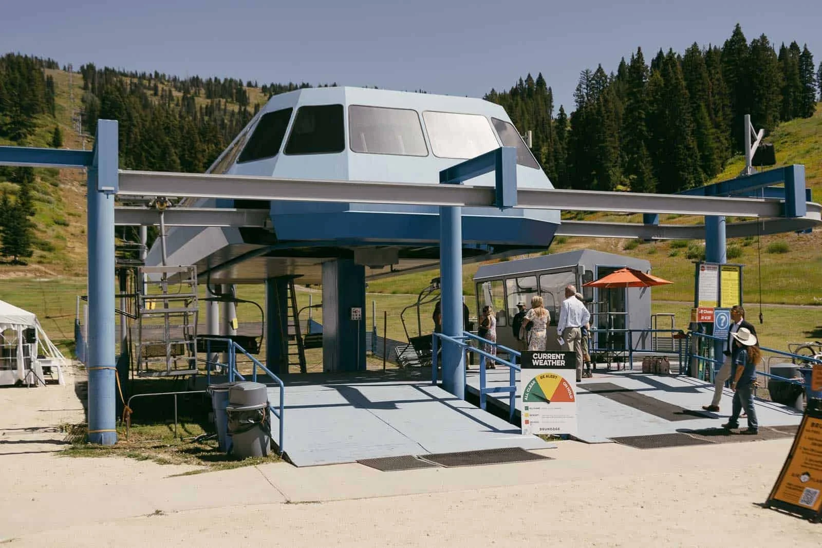  Wedding guests in summer dresses and suits line up at the Bluebird Chairlift loading station at Brundage Mountain in McCall, Idaho, with a green ski slope and pine trees behind them. 